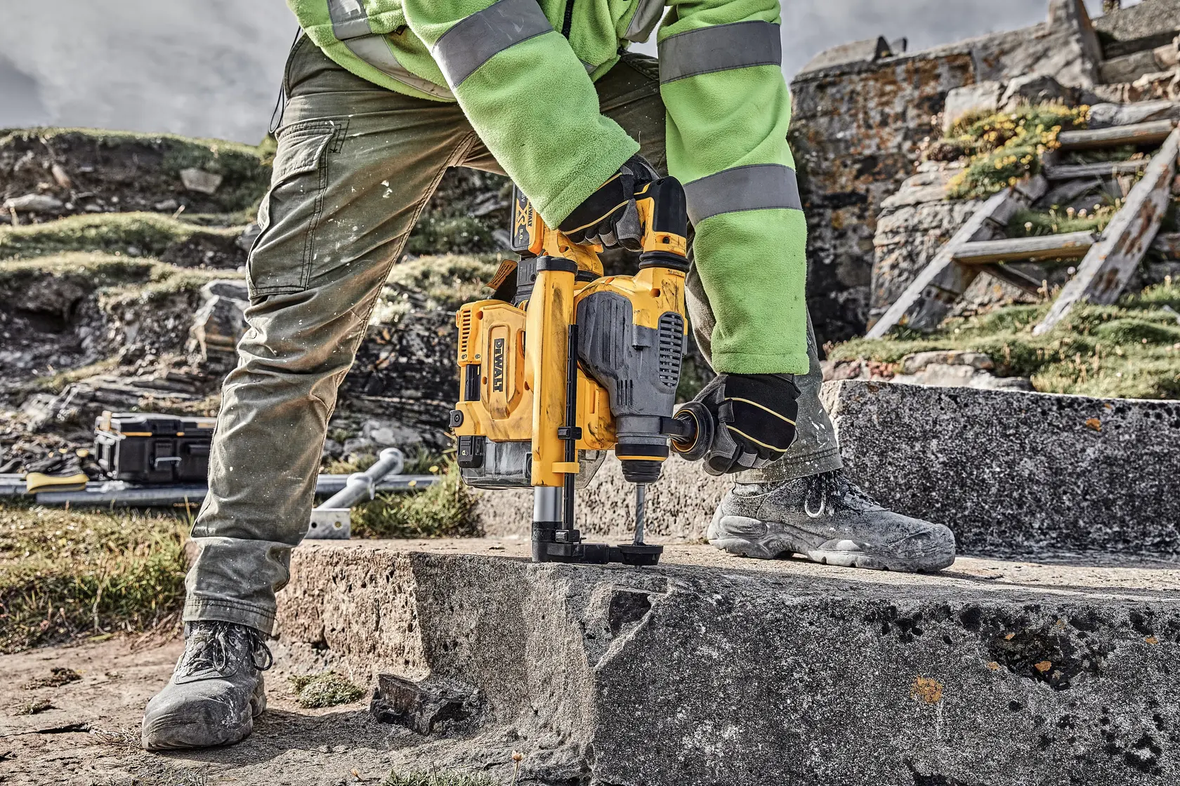 Person using DEWALT drill on concrete outdoors, wearing gloves, boots, and safety gear.
