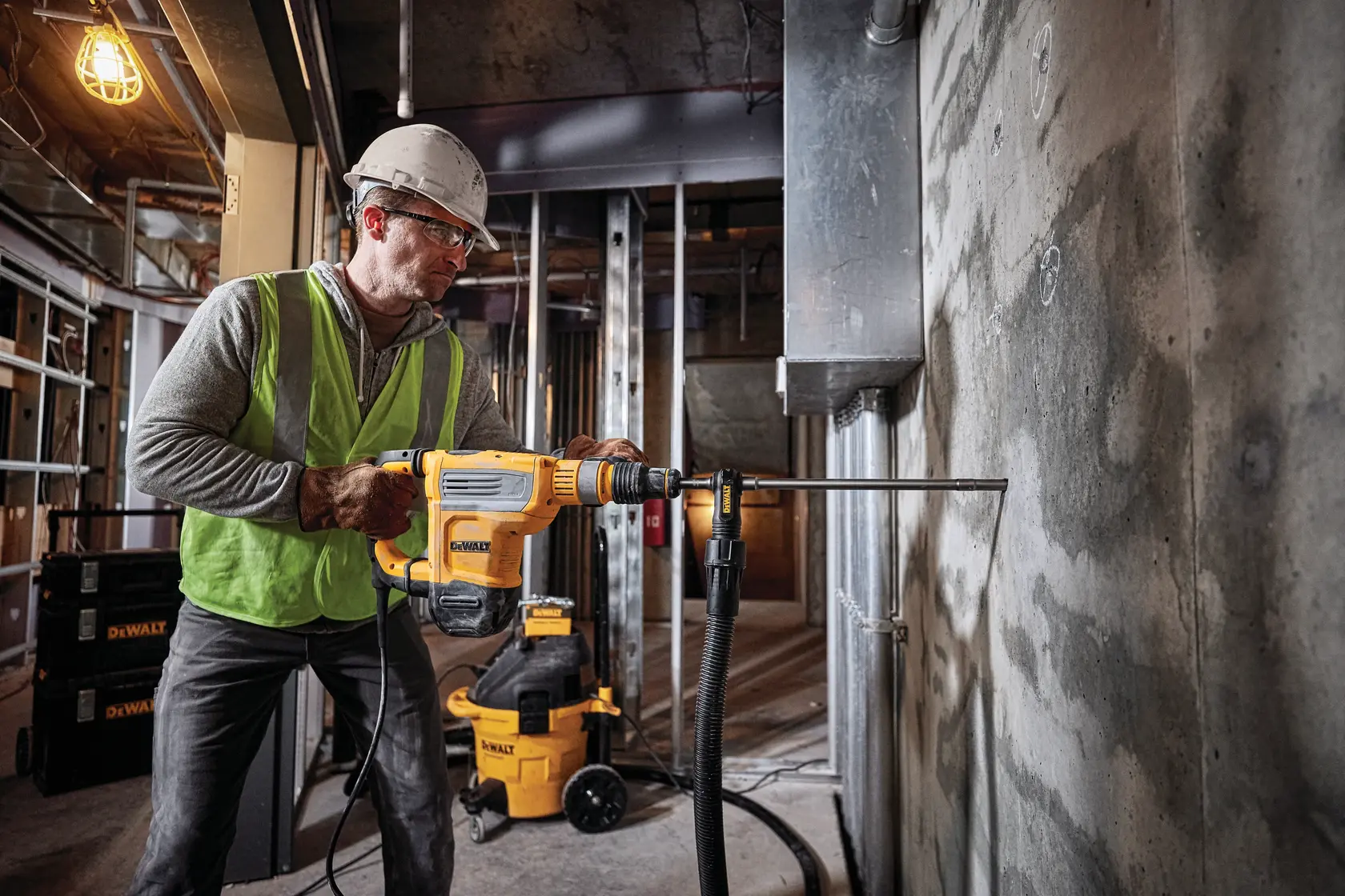 A person wearing safety gear and a helmet operates a DEWALT D25614K rotary hammer drill against a concrete wall in a construction site. DEWALT equipment and tools are visible in the background.