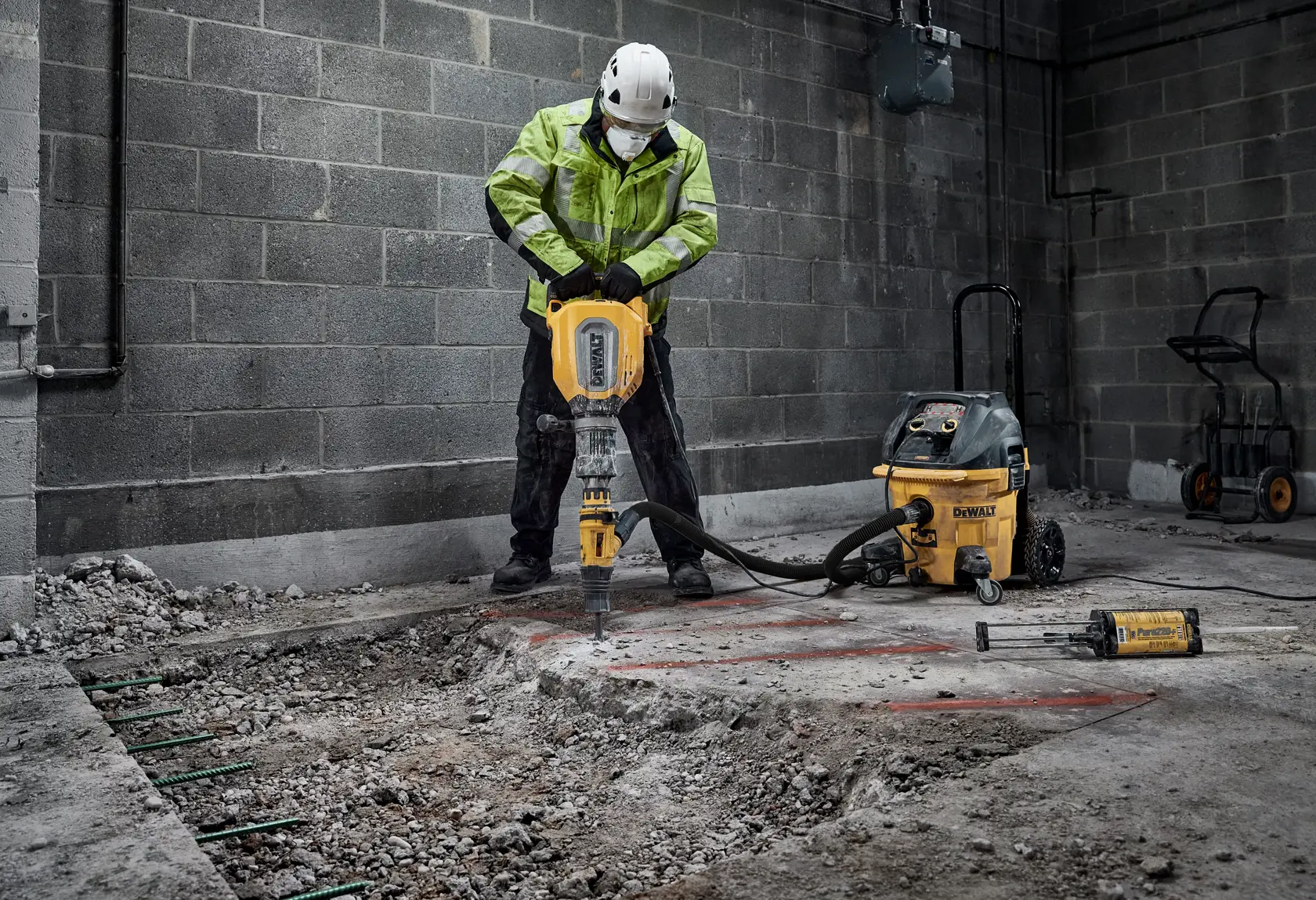 Worker using DEWALT demolition hammer to break concrete indoors. Safety gear and equipment visible.