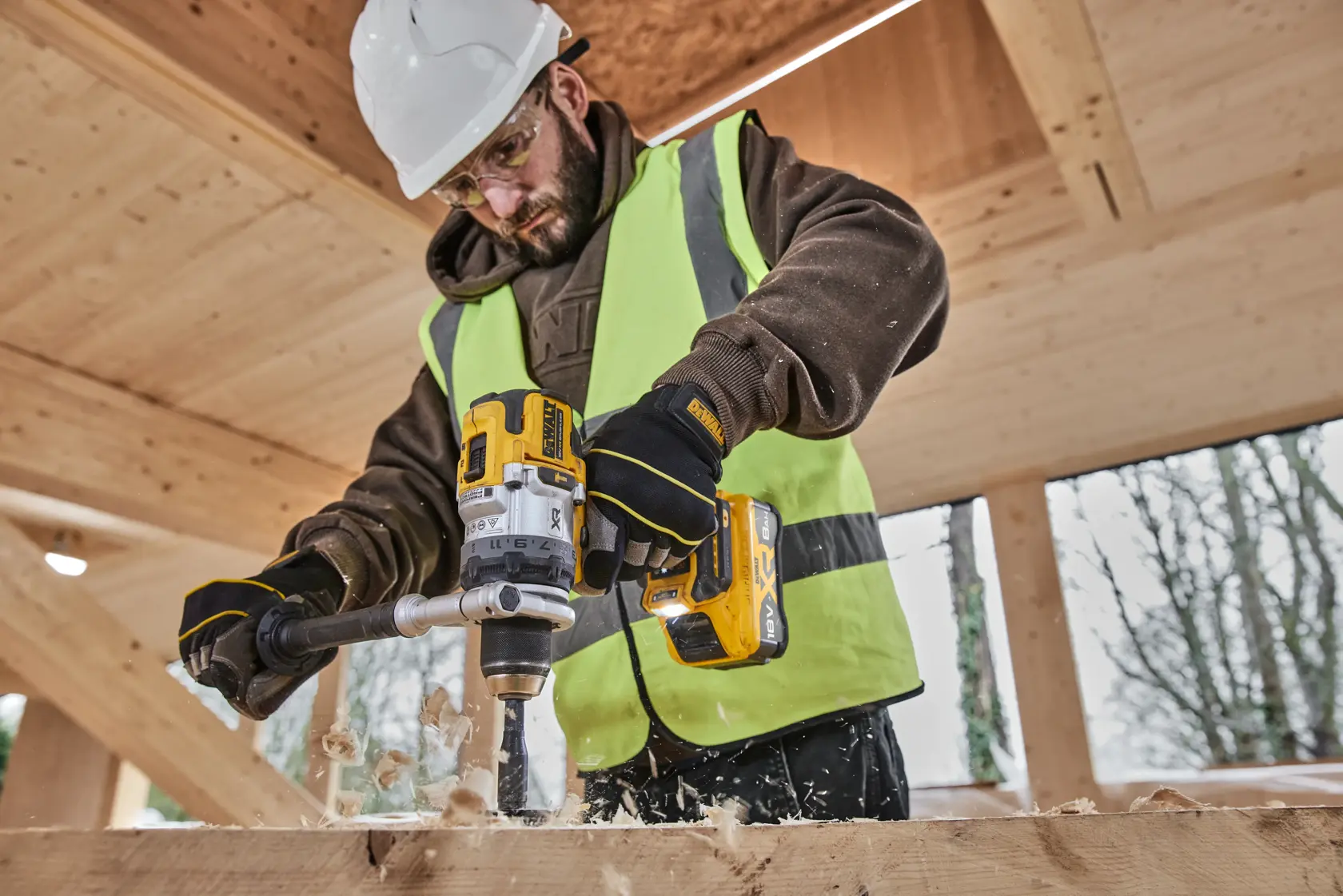 Person uses DEWALT drill with DCB1880 battery to drill wood. Wearing safety vest, gloves, and helmet at construction site.