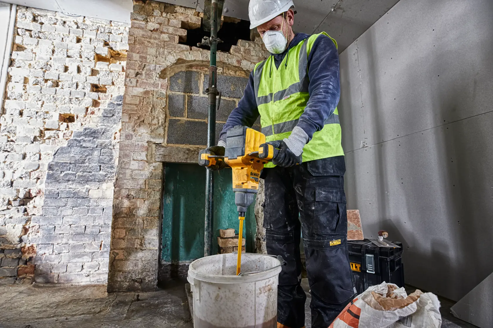 A person wearing a safety vest and gloves is using a DEWALT DCD240X2 cordless mixer to stir contents in a large bucket at a construction site with brick and drywall walls.