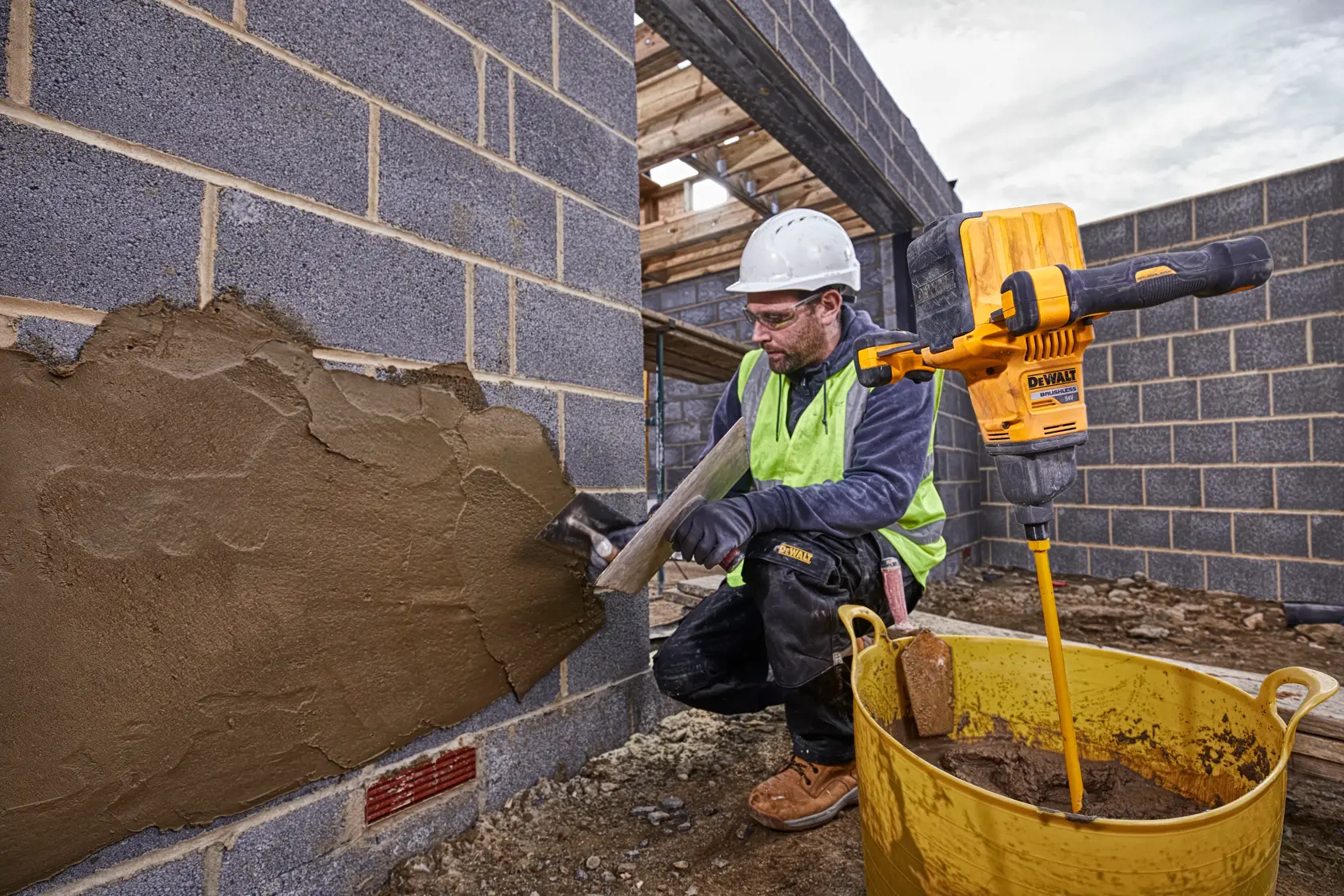 A construction worker wearing a safety helmet and a high-visibility vest applies plaster to a brick wall at a building site. Next to the worker is a yellow DEWALT mixing drill (model DCD240X2) placed in a large yellow container filled with plaster mix.