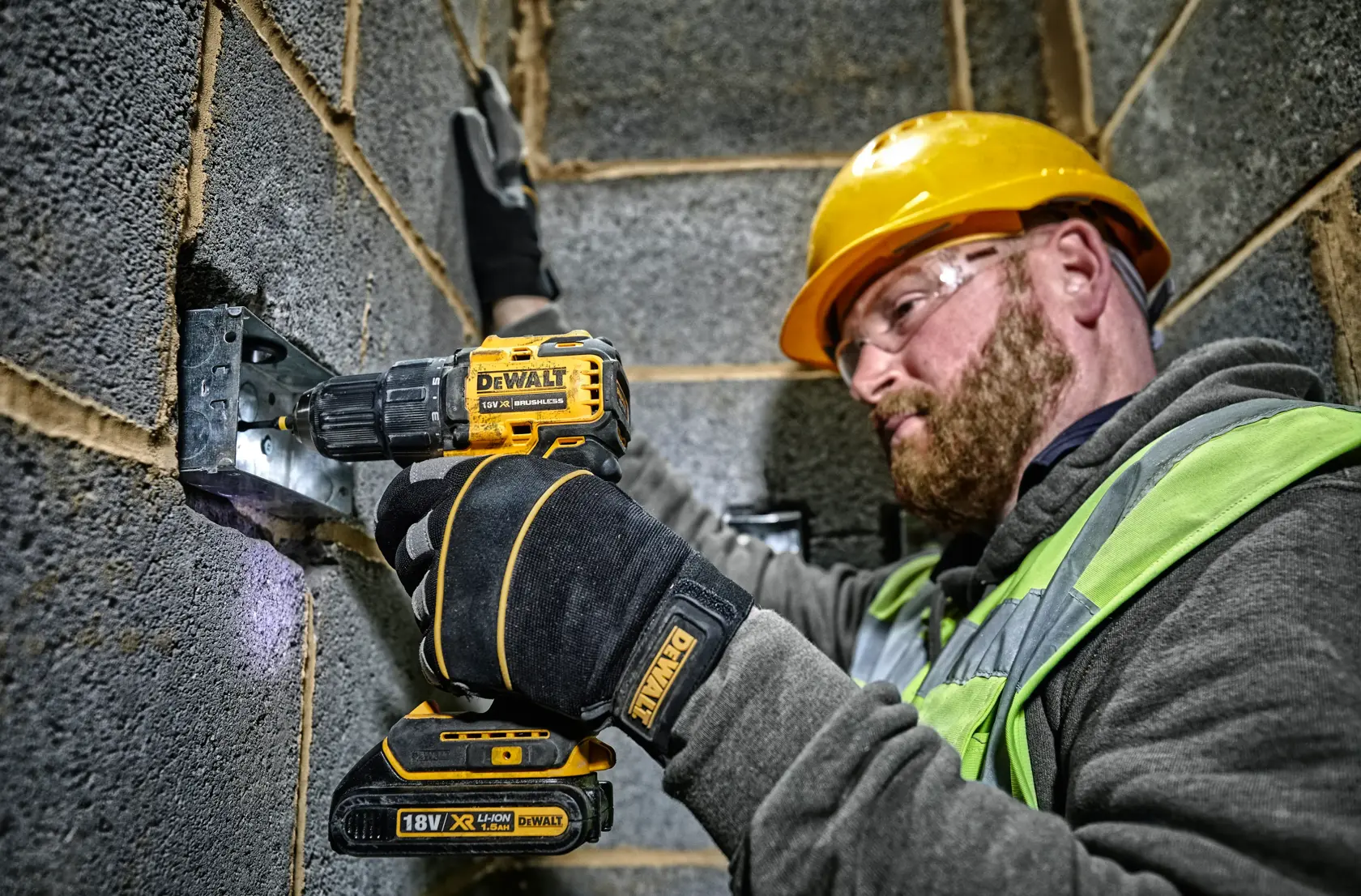A construction worker wearing a yellow hard hat and gloves is using a DEWALT DCD708 cordless drill to fasten a metal bracket to a cinder block wall. The worker’s face is blurred and he is dressed in a high-visibility vest and grey hoodie.