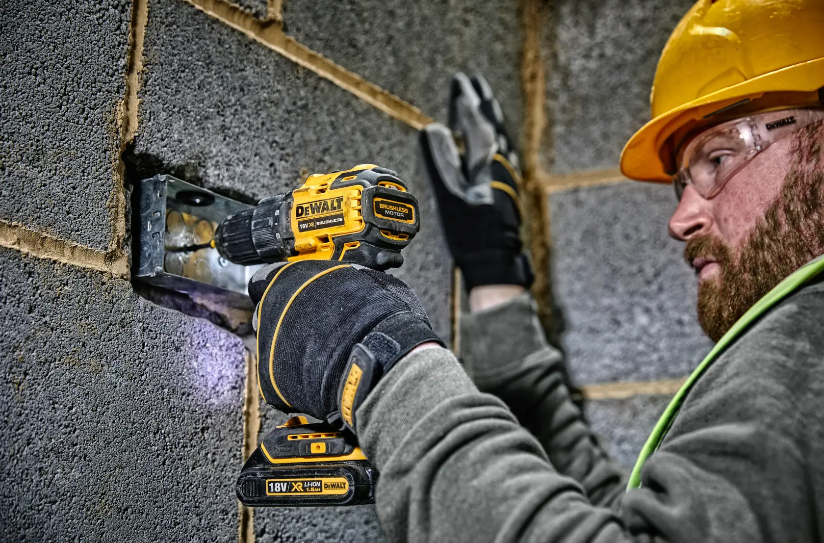 A person wearing gloves and a safety helmet is using a DEWALT 18V cordless drill driver (model DCD708) to work on a metal fixture in a cinder block wall.