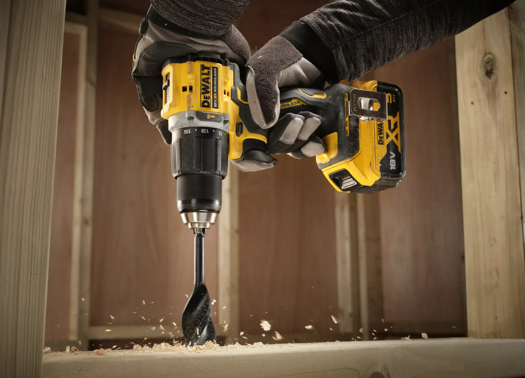 Gloved hands using a DEWALT cordless drill to bore into wood, producing shavings as it drills.
