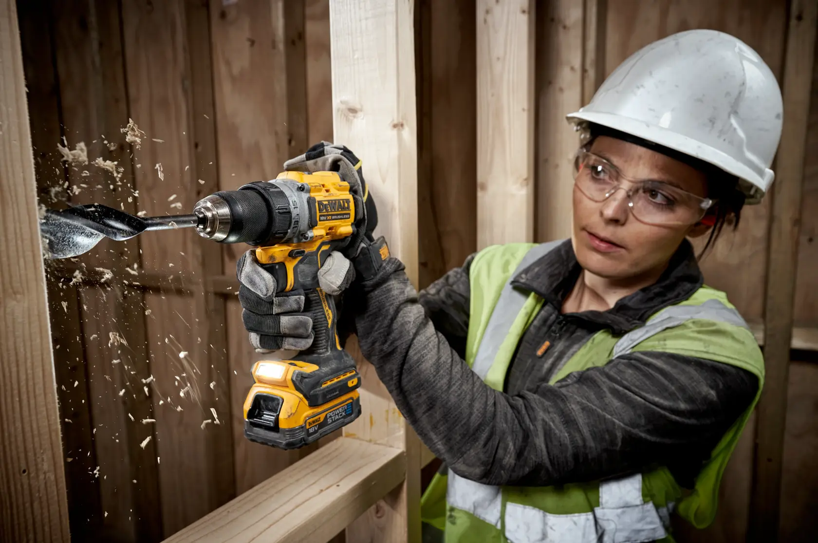 Person uses DEWALT cordless drill to bore into wood, wearing safety gear at a construction site.