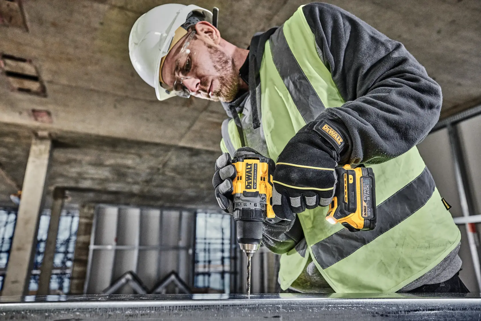 A person wearing a safety vest, gloves, and a hard hat is using a DEWALT DCD800NT cordless drill to drill into a metal surface at a construction site.