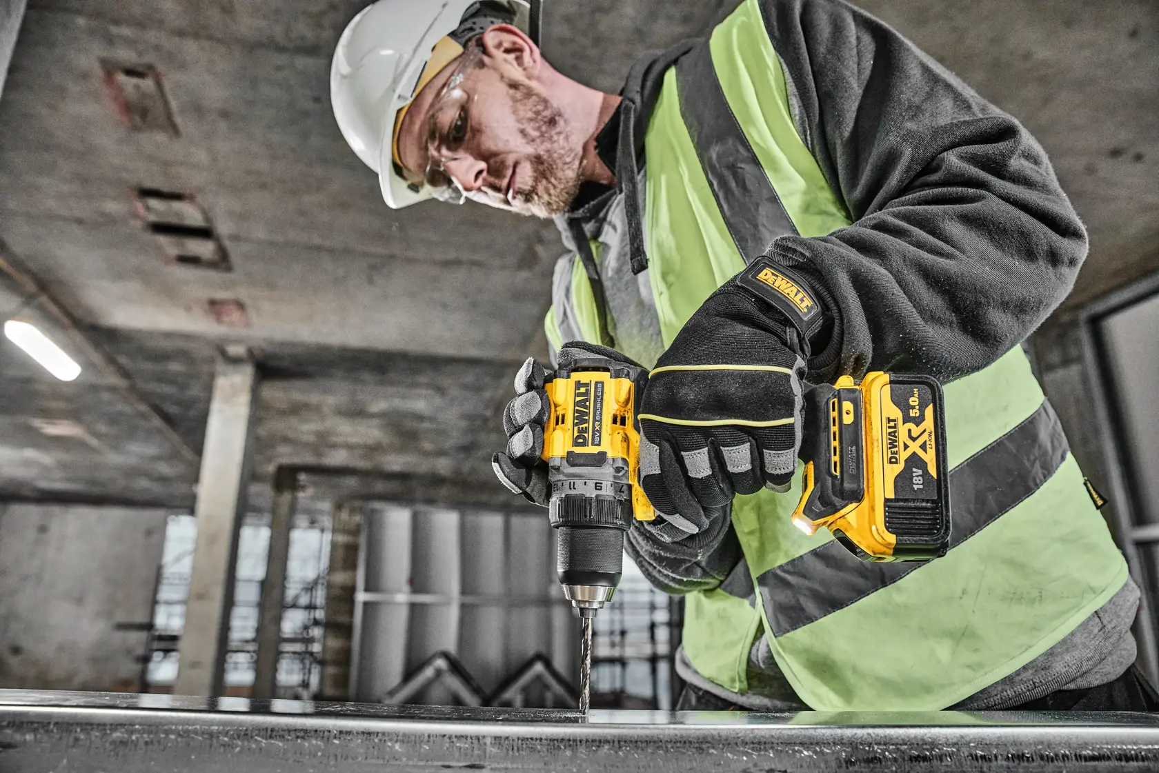 A person wearing safety gear, including gloves, a helmet, and a hi-vis vest, is using a DEWALT DCD800P2T cordless drill to work on a metal surface in an industrial environment.