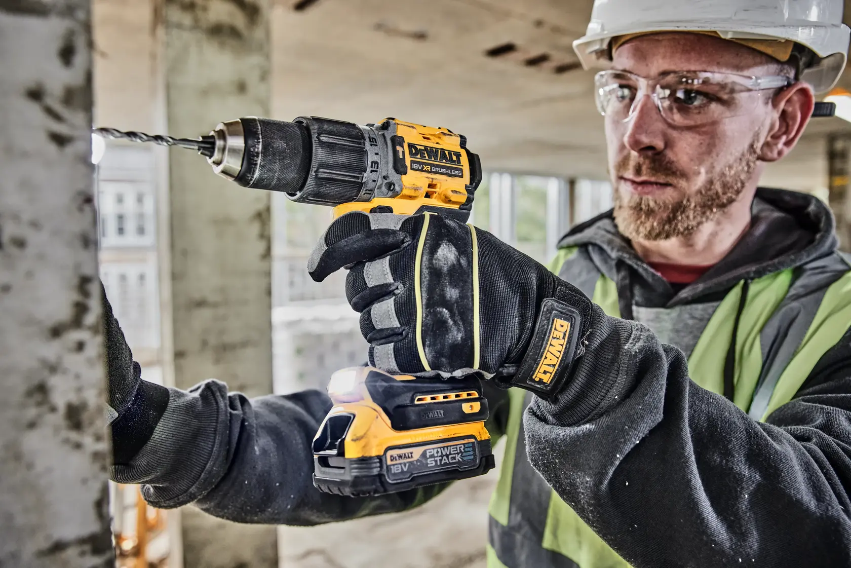 A person wearing work gloves and safety gear is drilling into a metal surface using a yellow and black DEWALT cordless power drill at a construction site.