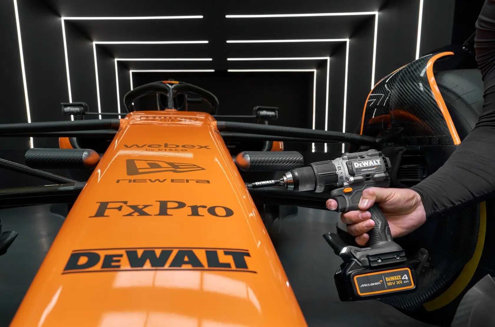 DEWALT drill held near an orange race car in a modern, illuminated garage with visible sponsor logos.