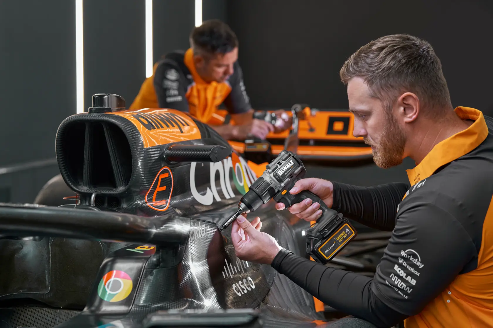 Person uses a DEWALT drill on a race car in a workshop. Faces are blurred. DEWALT branding is visible.