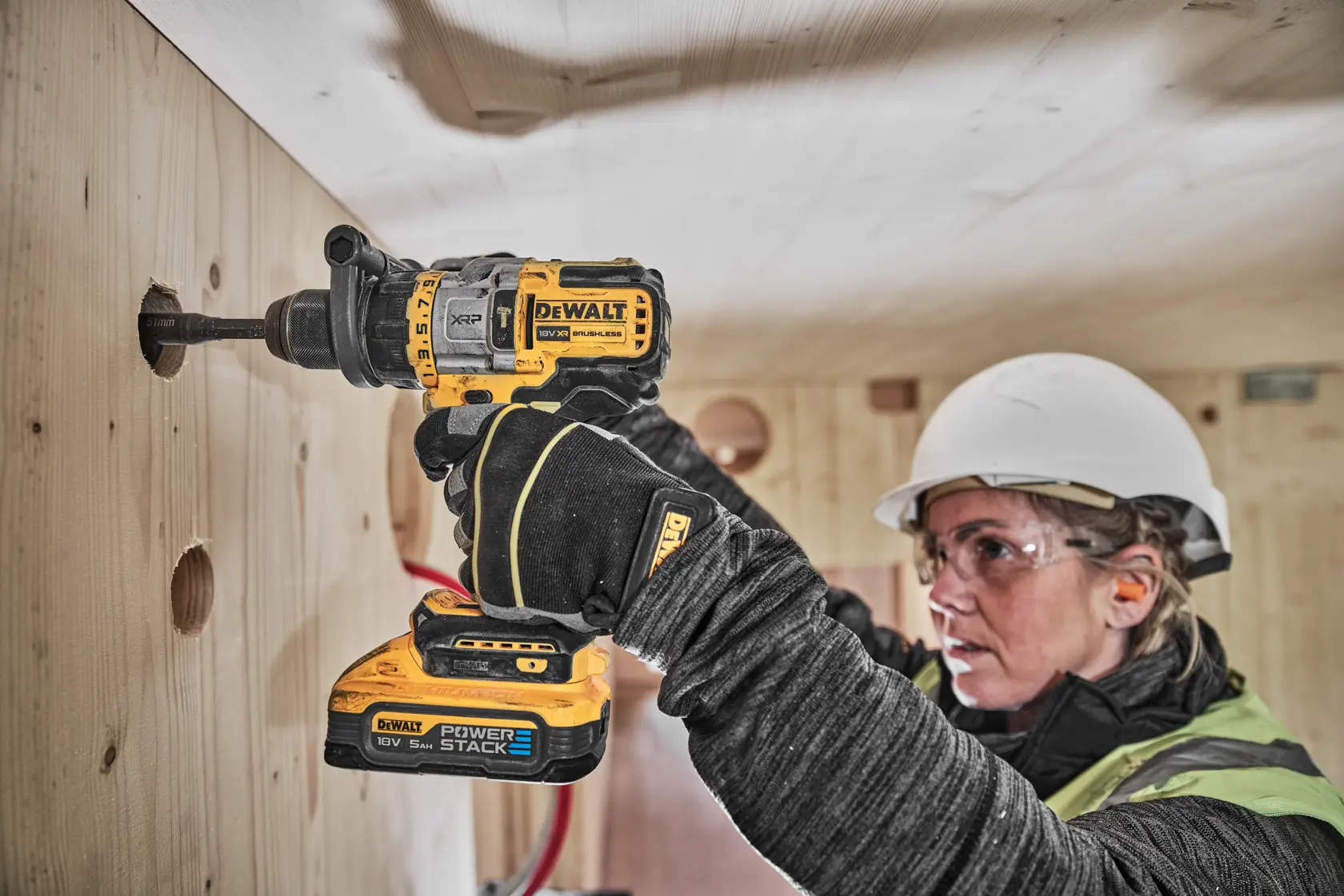 A person wearing safety gear is using a DEWALT DCD999H2T cordless drill to bore a hole into a wooden wall. The drill is fitted with a POWERSTACK battery.