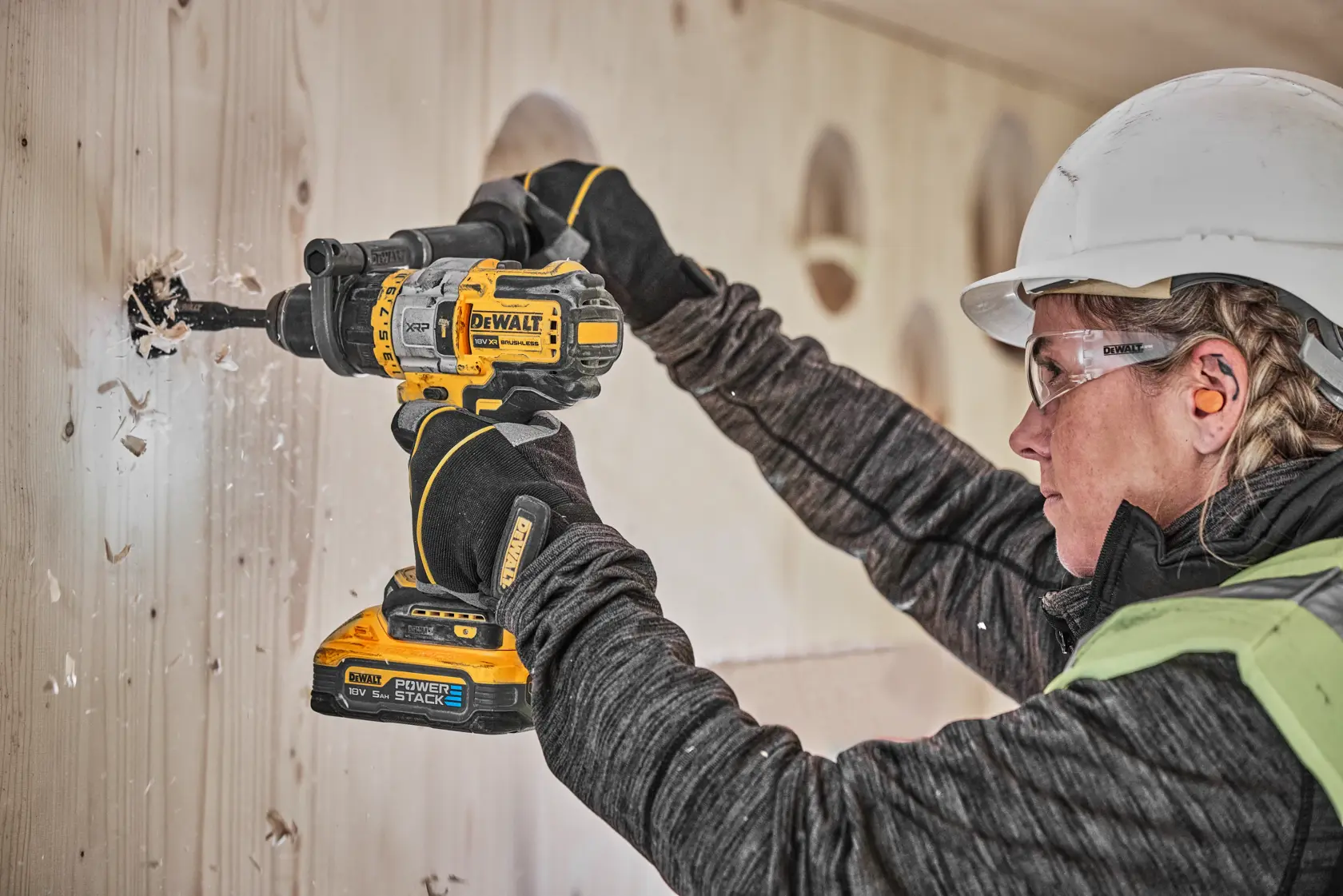 A person wearing safety gear uses a DEWALT DCD999H2T cordless drill to drill a hole in a wooden wall.