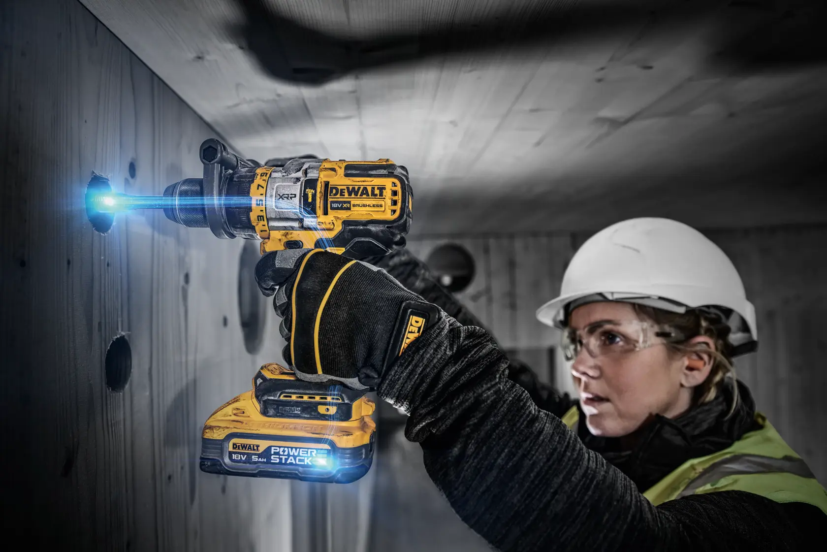 A person wearing safety gear uses a DEWALT DCD999H2T PowerStack cordless drill to bore a hole into a wooden wall. The drill is illuminated, and the person's face is blurred for privacy.