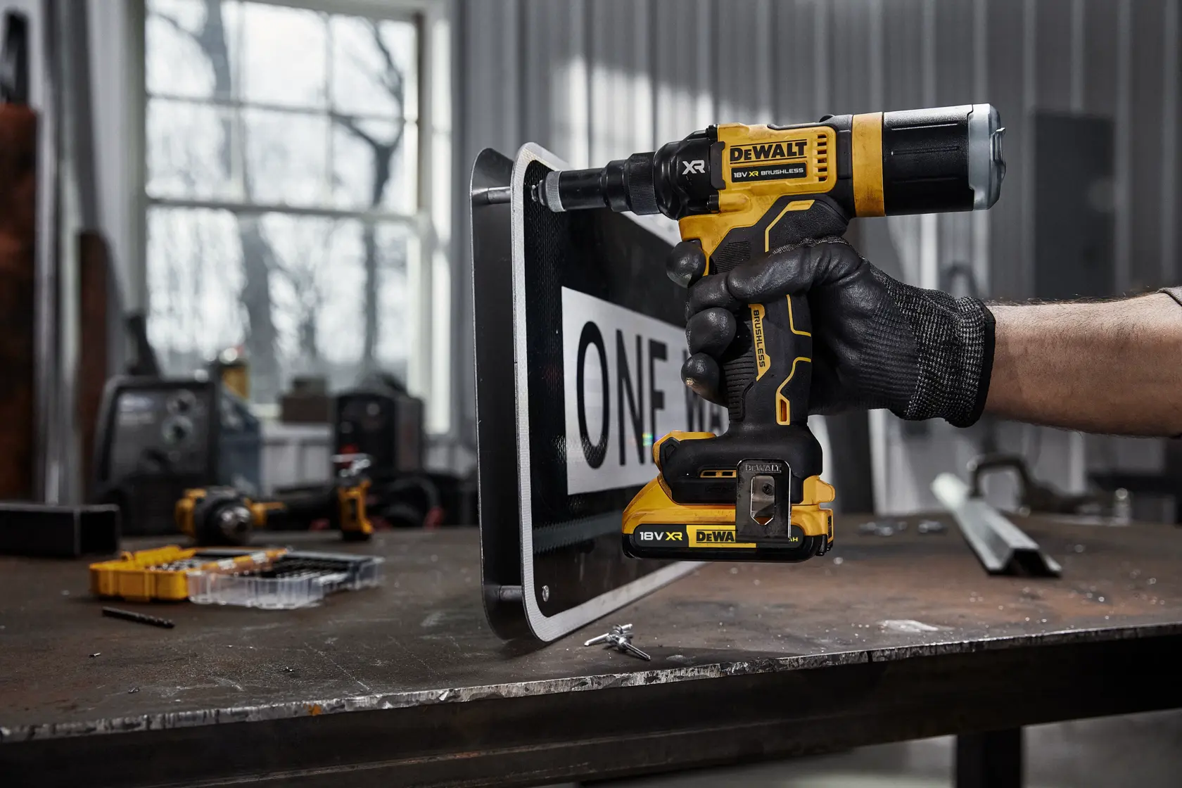 A person wearing a black work glove uses a DEWALT DCF403NT 18V XR cordless drill to drill into a metal sign on a workbench in a workshop.