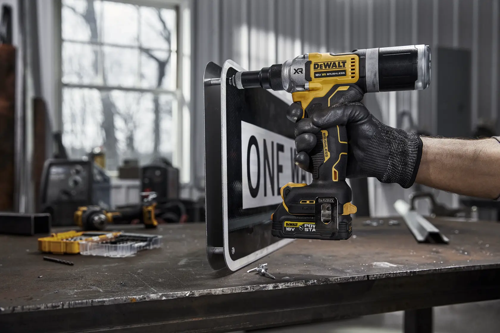 A person wearing a black glove is using a yellow and black DEWALT DCF414E2GT power tool to drill or fasten a street sign on a metal workbench in an industrial workshop.