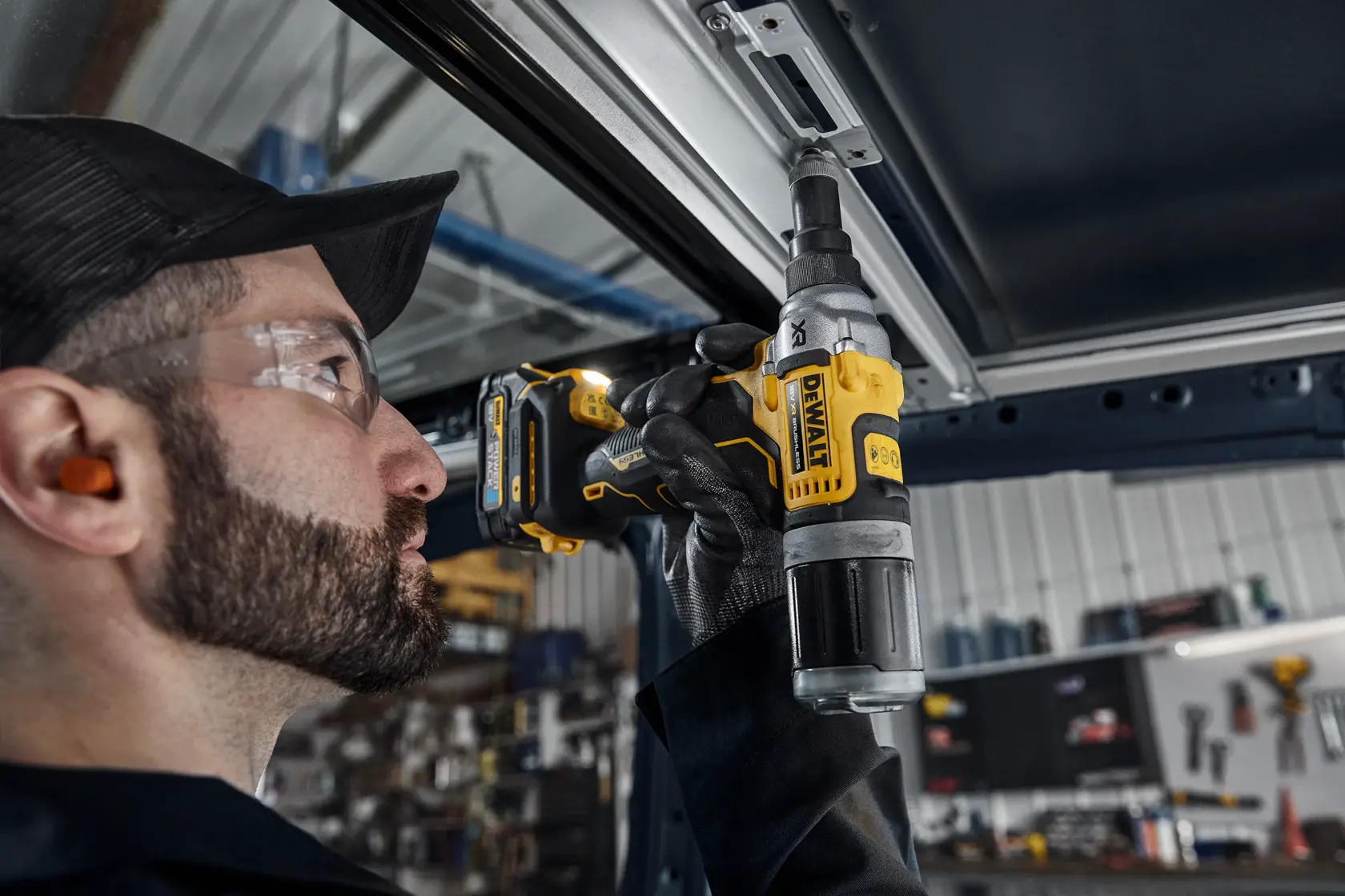 A person wearing ear protection and gloves uses a DEWALT DCF414N cordless power tool to fasten or unfasten a metal component indoors, in an industrial or workshop setting.