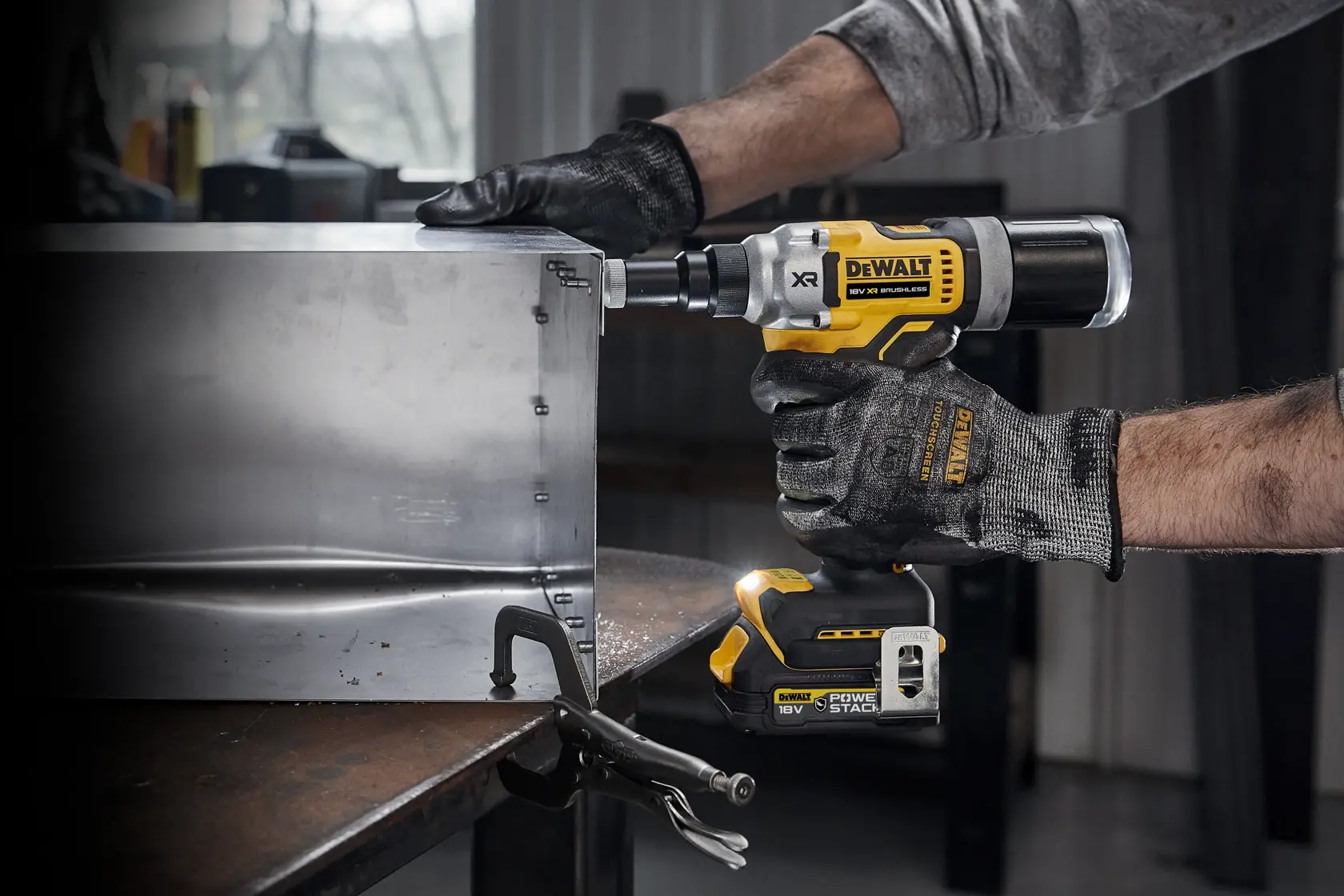A person wearing work gloves is using a DEWALT DCF414NT 18V cordless rivet gun to fasten a metal sheet in an industrial workshop.