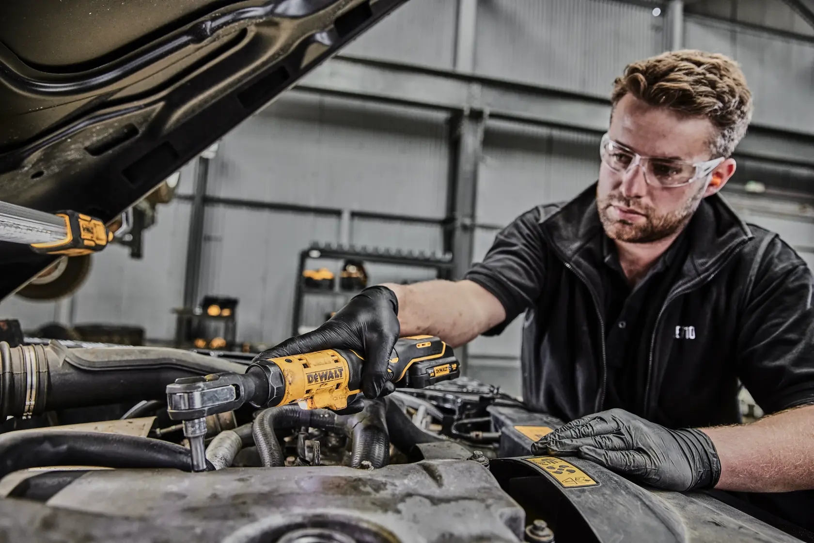 A person wearing black gloves uses a DEWALT cordless ratchet tool to work on a car engine in an industrial workshop. The person's face is blurred.