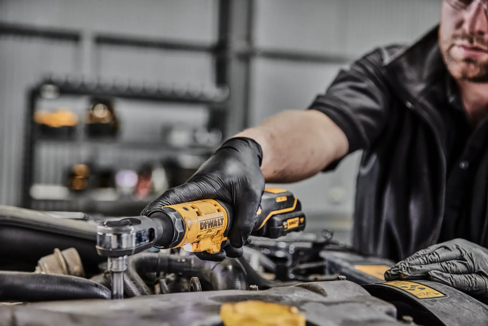 A person wearing black gloves uses a yellow DEWALT cordless ratchet tool (model DCF504L1G) to work on an engine in a workshop.