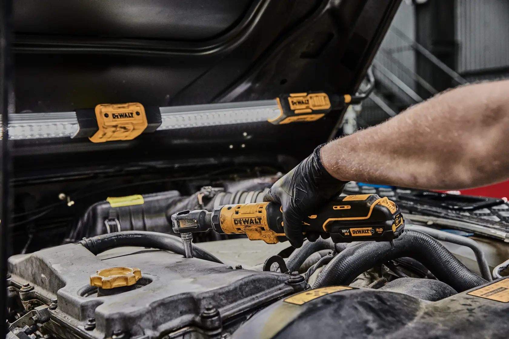 A person using a DEWALT cordless ratchet tool to work on a car engine, with a DEWALT work light attached to the underside of the car hood.