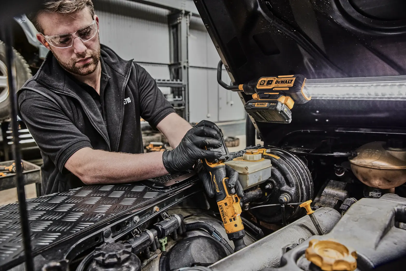 A person wearing black gloves uses a DEWALT power tool, likely a cordless ratchet, to work on the engine of a vehicle in a workshop. A DEWALT work light is attached to the underside of the open car hood, illuminating the work area.