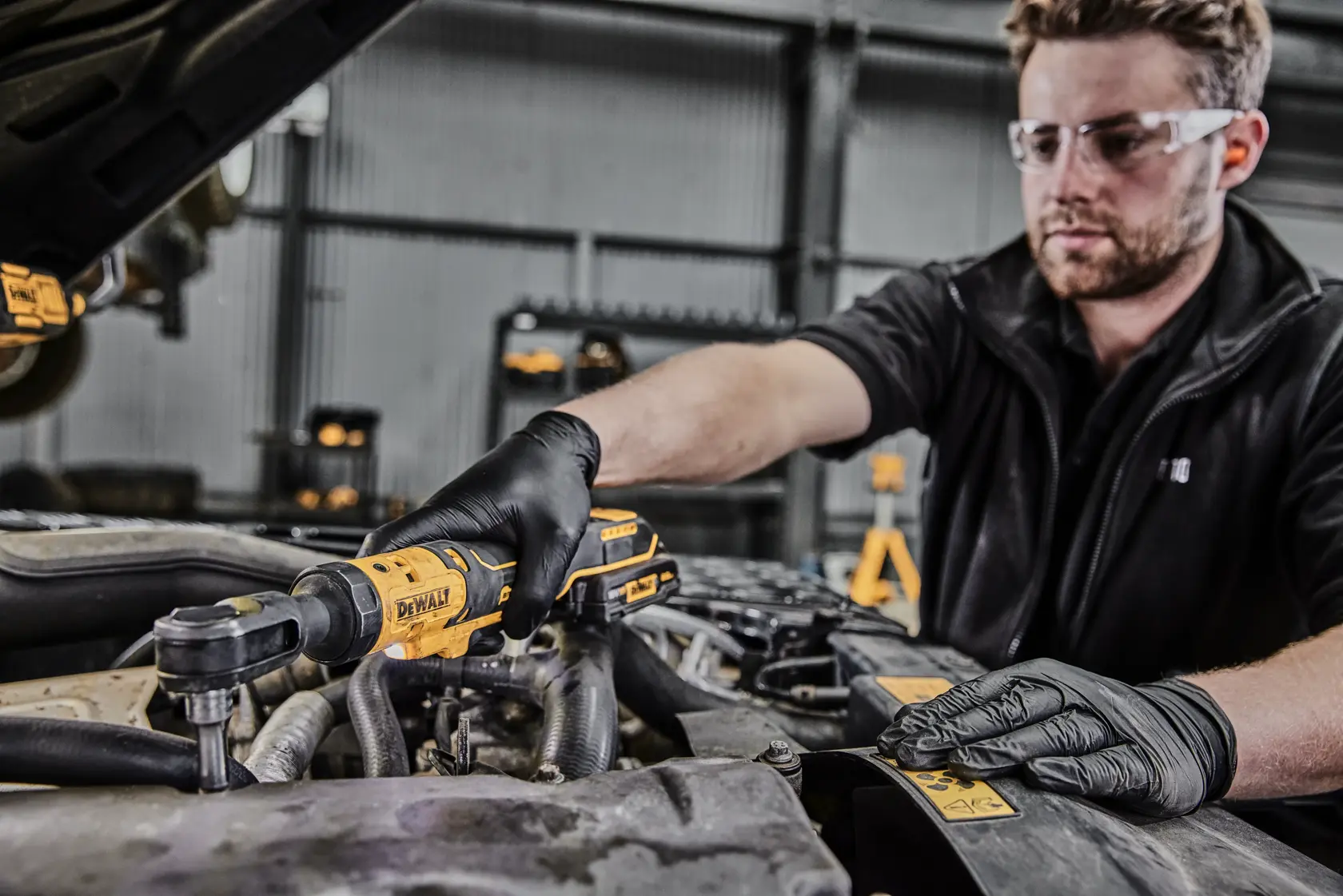Person wearing black gloves using a DEWALT DCF512 cordless ratchet wrench to work on a car engine in a workshop.