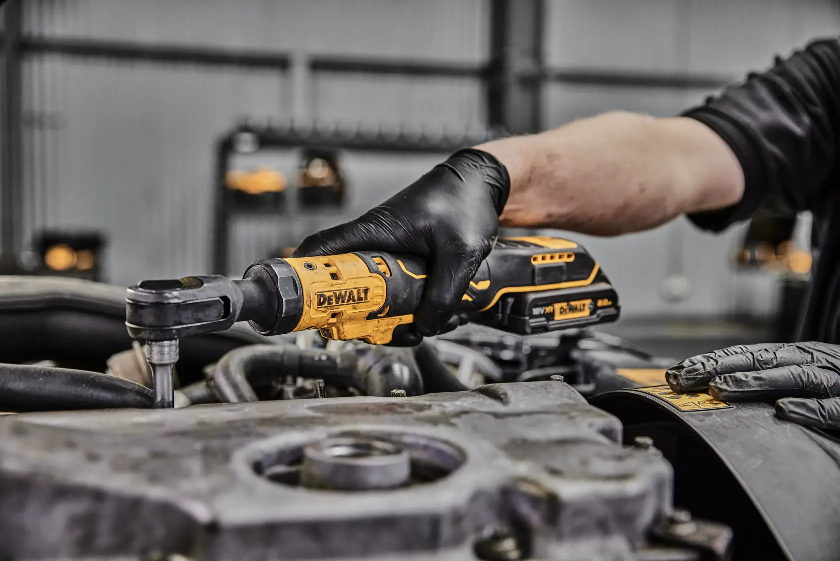 A person wearing black gloves uses a DEWALT cordless ratchet tool, model DCF512D1G, to work on a mechanical component, possibly in an automotive setting.