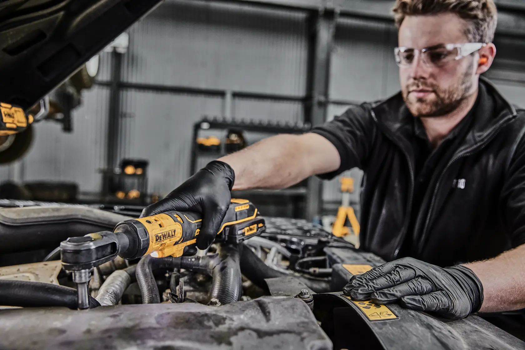 A person wearing black gloves uses a DEWALT DCF512N cordless ratchet tool to work on the engine of a vehicle. Their face is