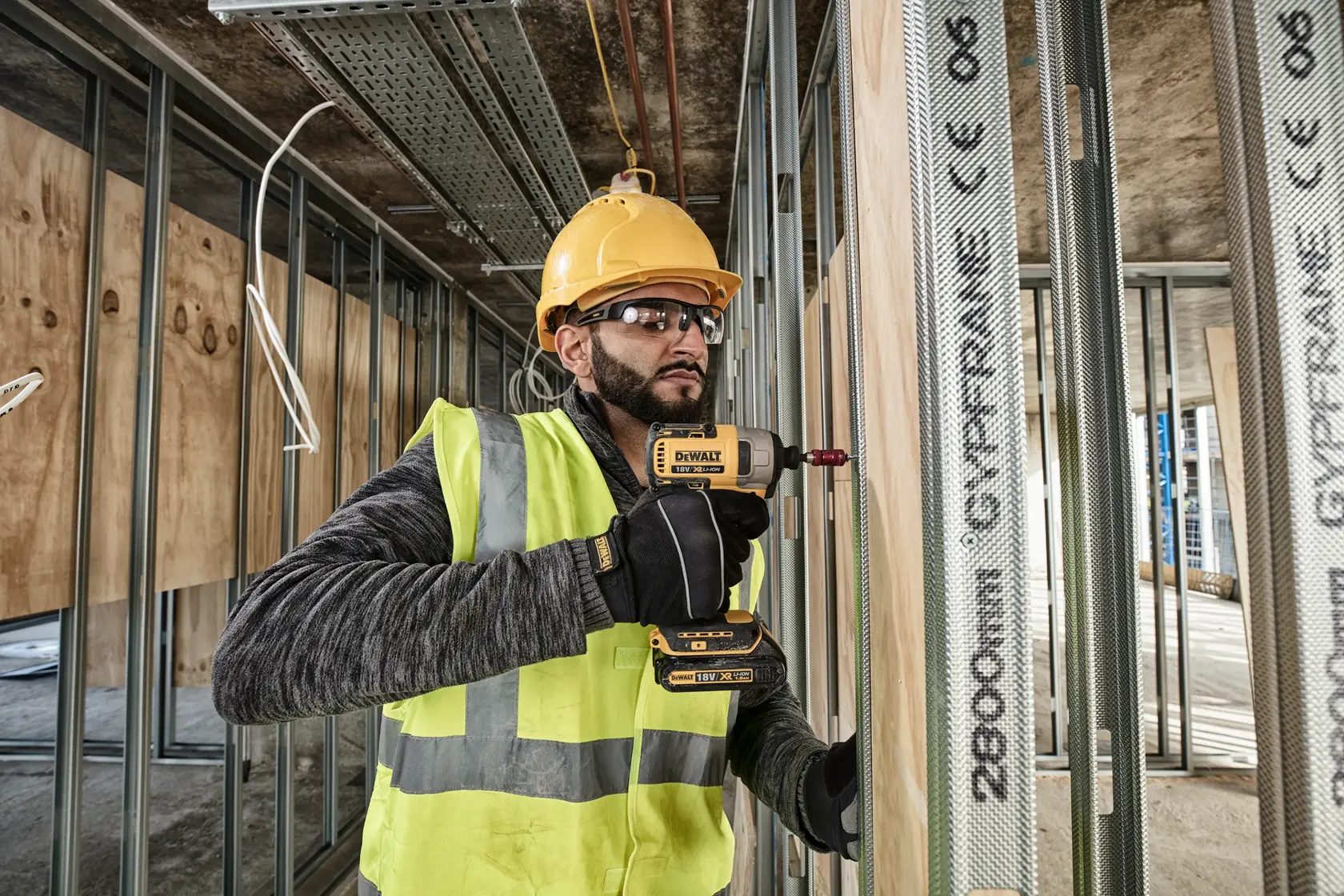 Worker using DEWALT DCF787N drill on metal studs at construction site, wearing safety gear and yellow helmet.