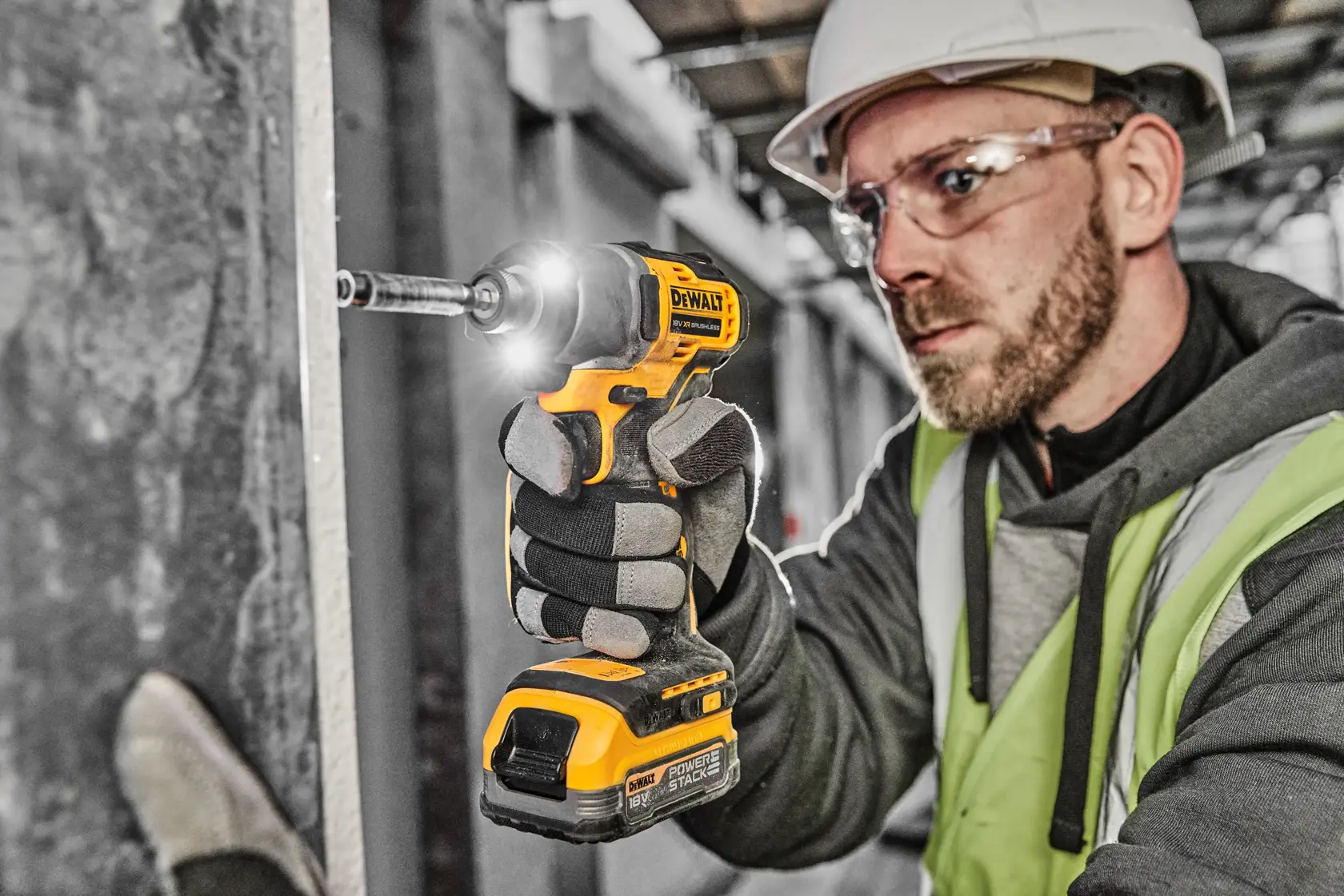 A person wearing safety gear uses a DEWALT DCF840NT cordless impact driver to fasten a screw into a wall at a construction site. The tool is yellow and black, and the person’s face is obscured.