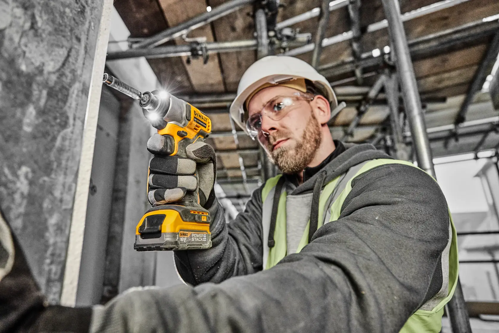 A person wearing safety gear uses a yellow DEWALT cordless drill to fasten a screw into a metal surface on a construction site.