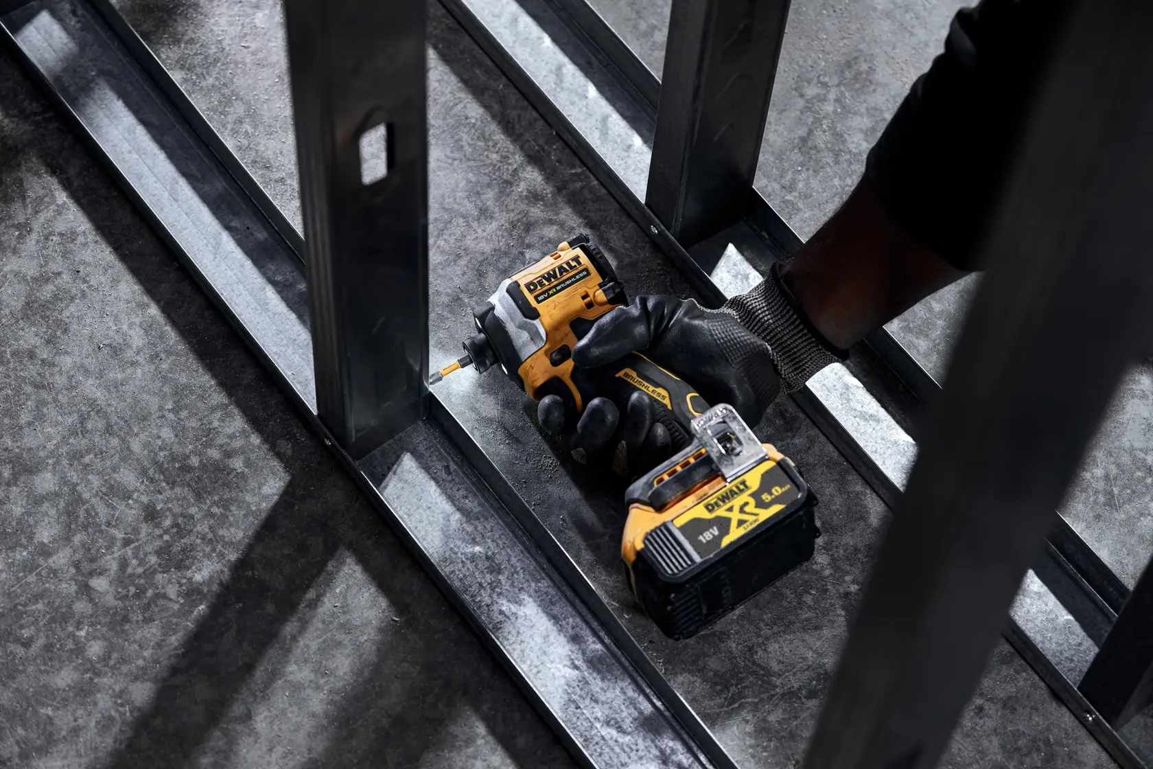 Gloved hand using a DEWALT cordless impact driver on steel framing indoors.
