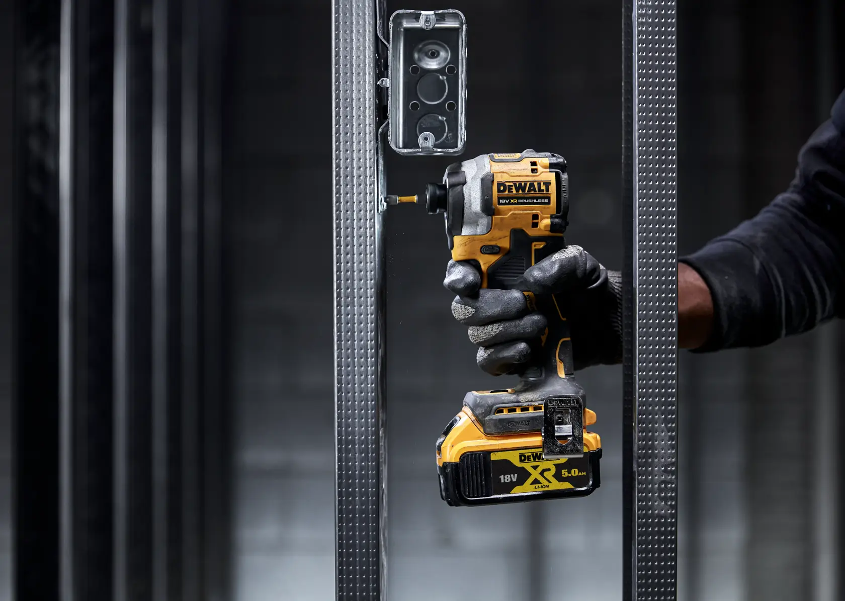 Gloved hand using a DEWALT cordless impact driver on a metal frame near an electrical box.