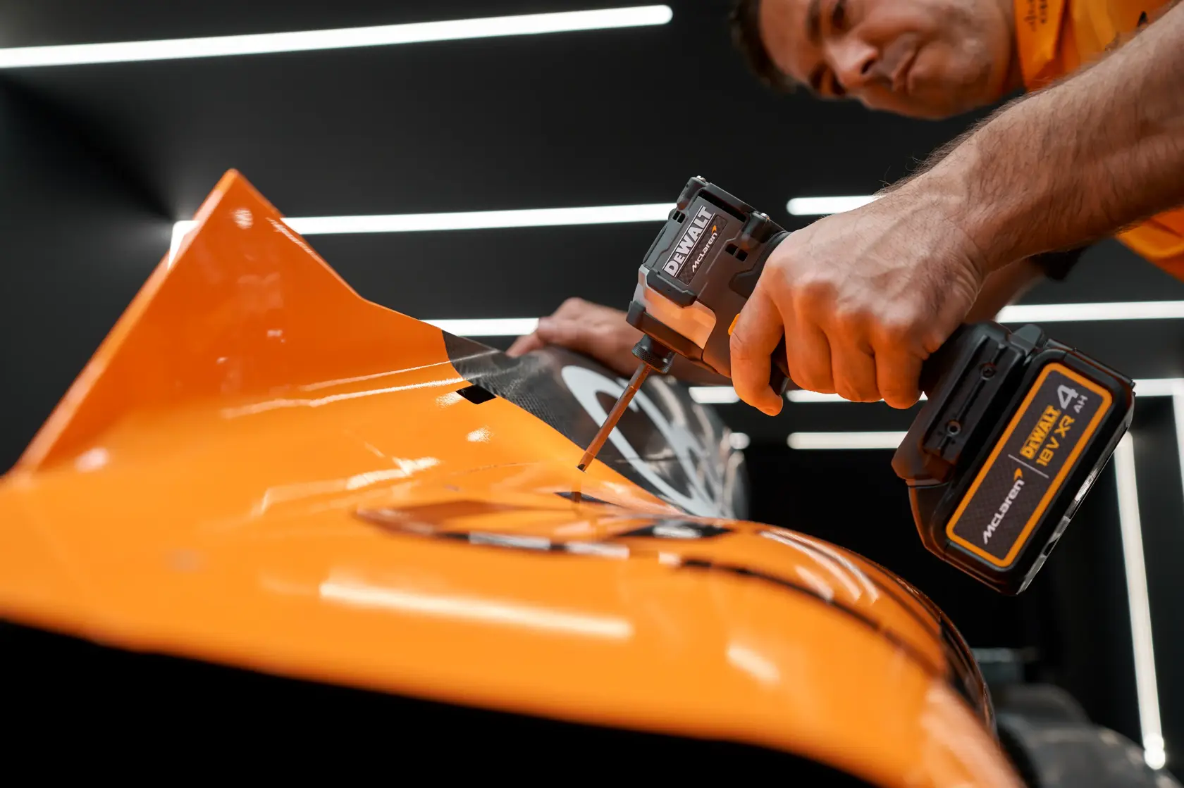 Person uses DEWALT cordless tool to fasten screws on an orange vehicle part in a modern workshop.