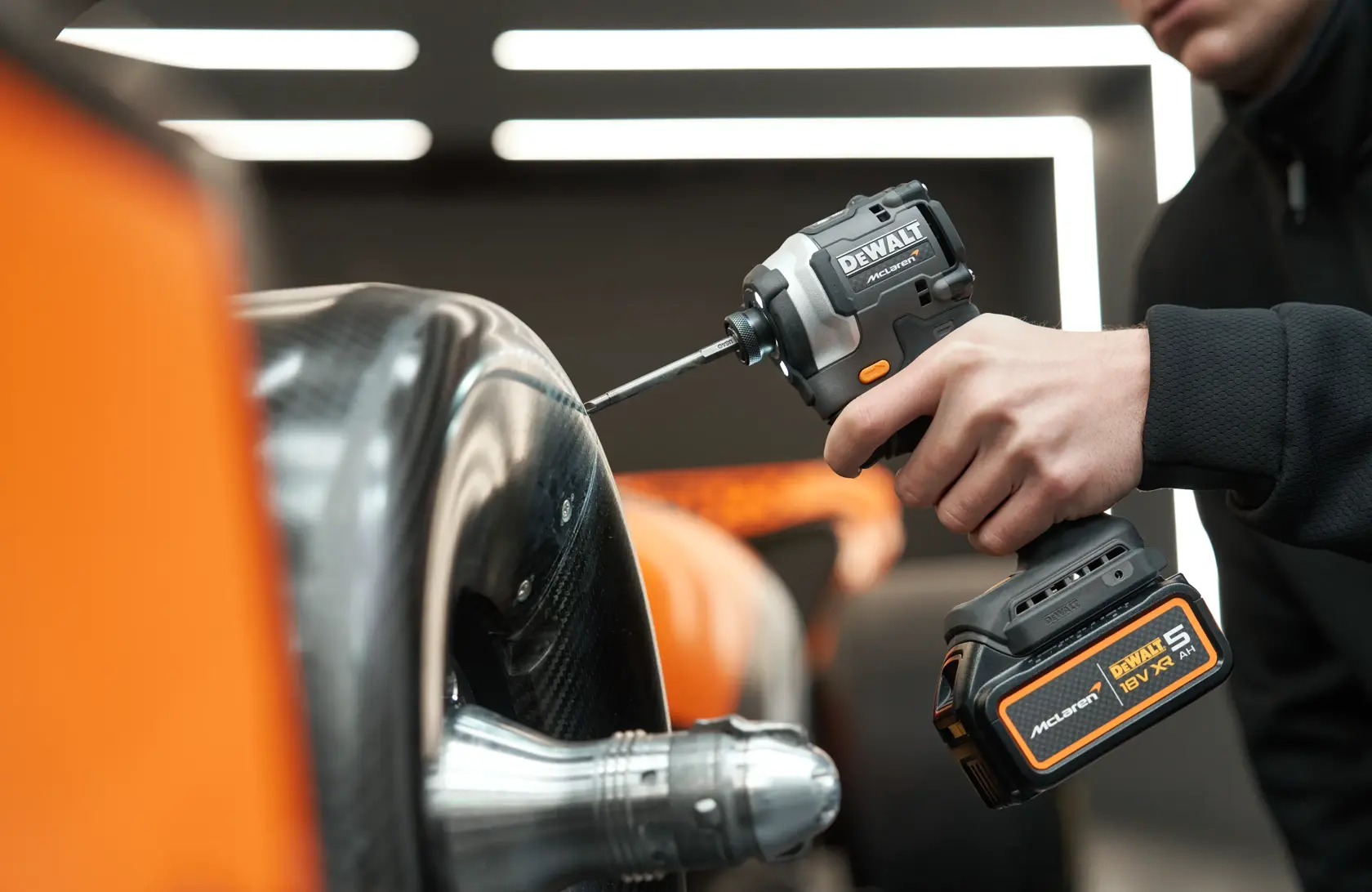 Person uses a DEWALT power tool to work on a car part in an indoor setting with orange and black details.