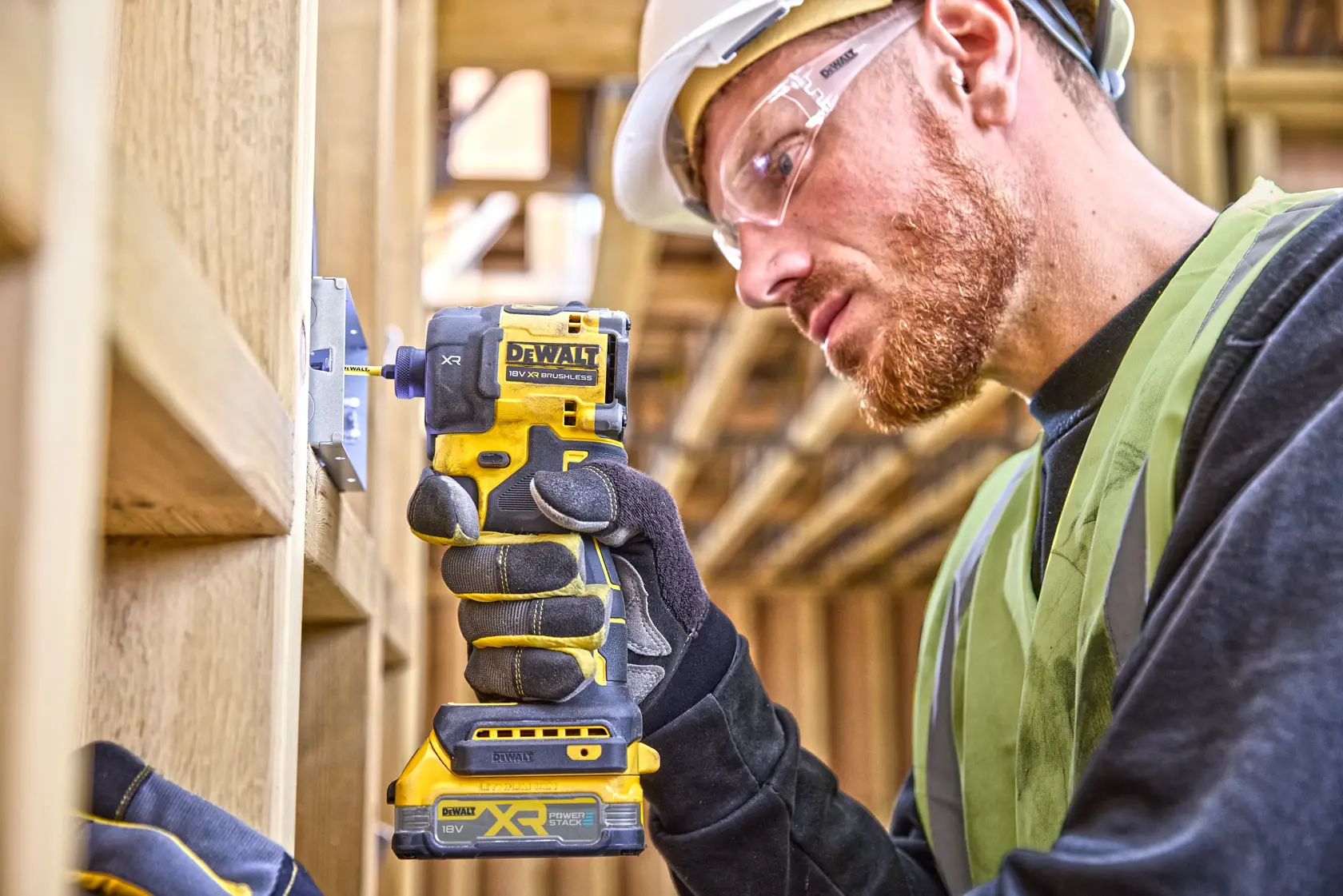 Worker uses DEWALT DCF870E2T impact driver on a wood frame, wearing gloves, helmet, and safety vest.
