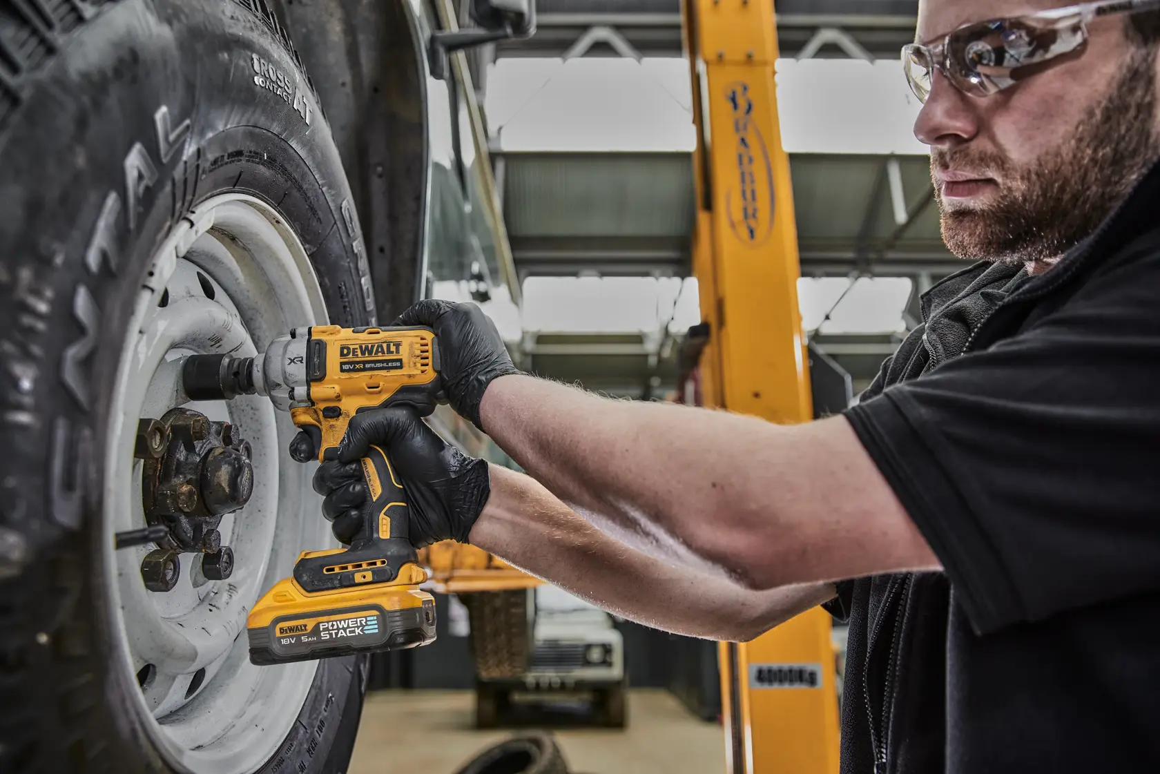 A person wearing black gloves uses a DEWALT DCF891H2T cordless impact wrench to tighten or loosen the lug nuts on a vehicle wheel in a garage setting.