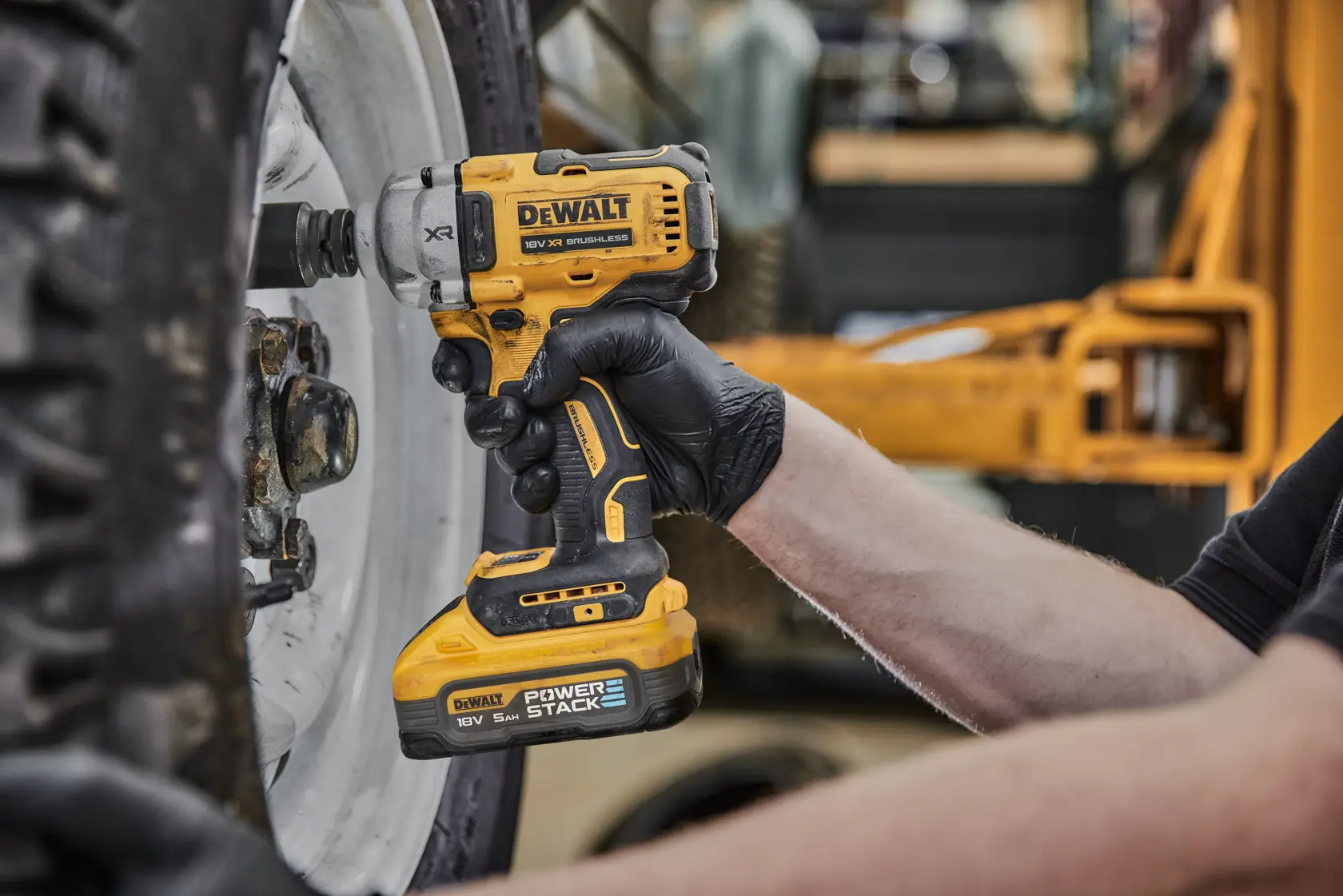A person using a DEWALT DCF891H2T cordless impact wrench to loosen bolts on a large vehicle wheel. The tool is yellow and black, and the user is wearing black gloves.