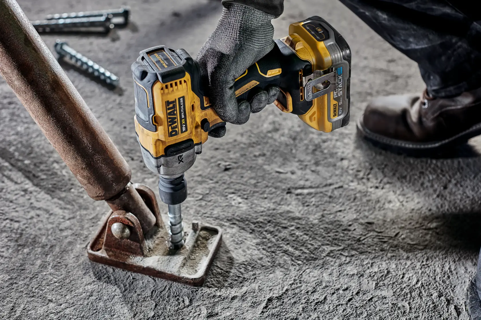 A person wearing work gloves and boots using a DEWALT cordless impact wrench to tighten a bolt on a rusty metal bracket attached to a concrete surface.