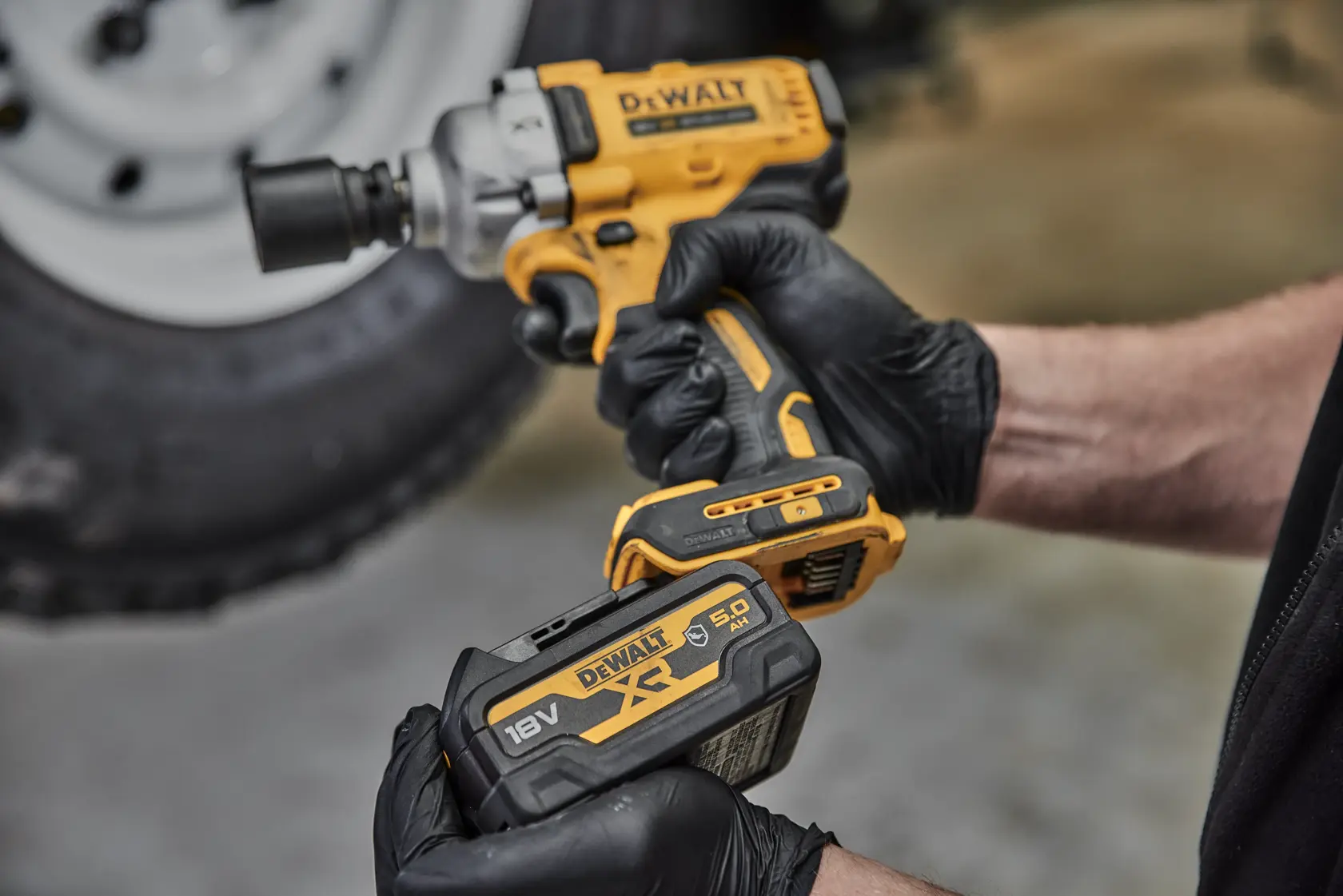 Gloved hands hold a DEWALT cordless impact wrench and an 18V battery in front of a blurred tire.