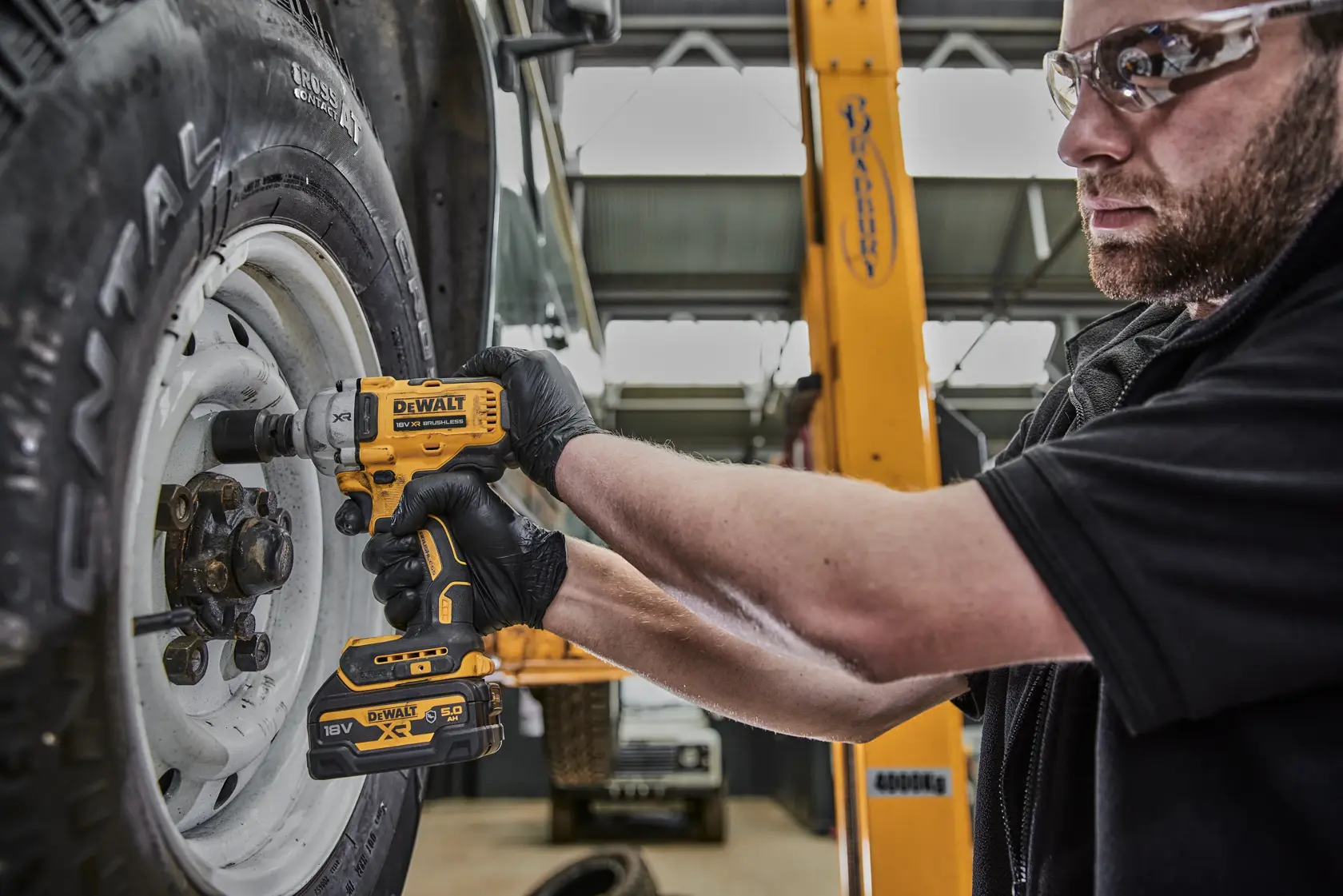 A person wearing black gloves uses a DEWALT DCF891P2G cordless impact wrench to tighten bolts on a vehicle wheel in a workshop setting.