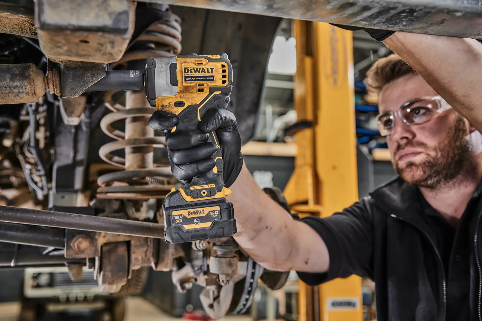 A person using a DEWALT cordless impact wrench (DCF901P1G) to work on the underside of a vehicle in a workshop. The person is wearing black gloves and the tool is being applied to a bolt.