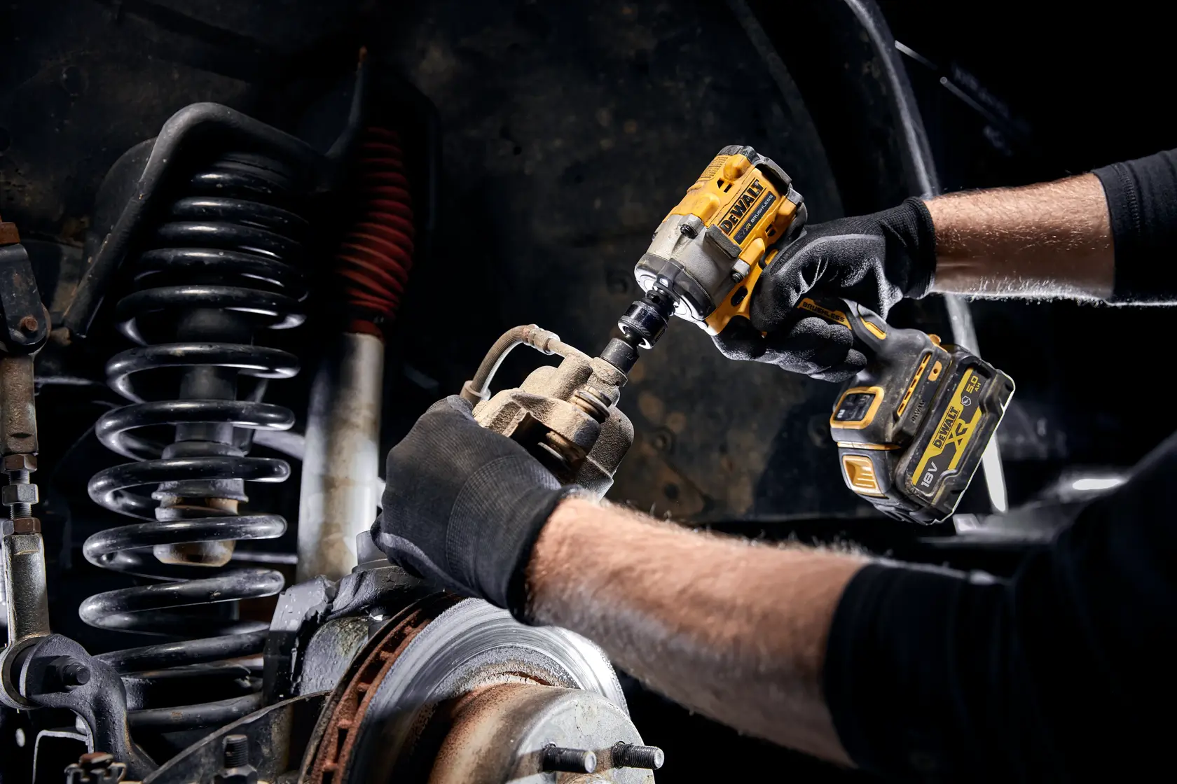 A person wearing black gloves uses a DEWALT cordless impact wrench to work on a vehicle's suspension and brake components.