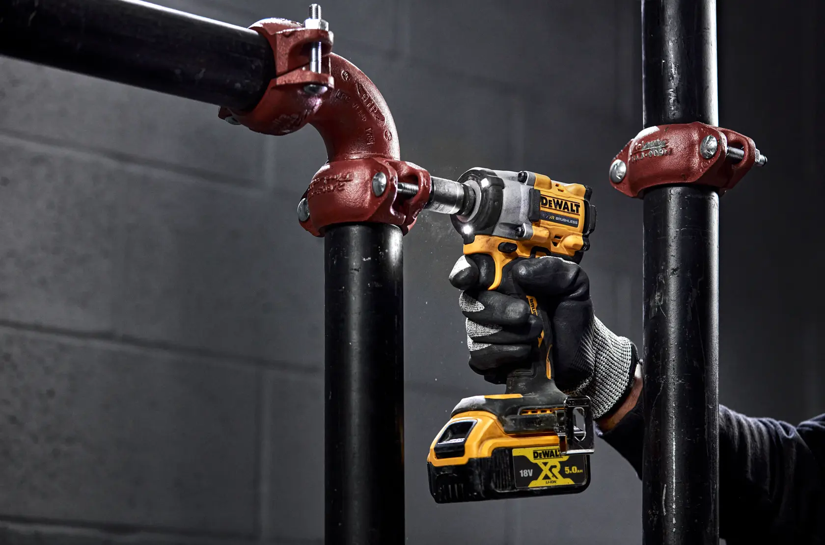 A person wearing black gloves uses a yellow and black DEWALT cordless impact wrench to tighten a bolt on a metal pipe fitting in an industrial setting.