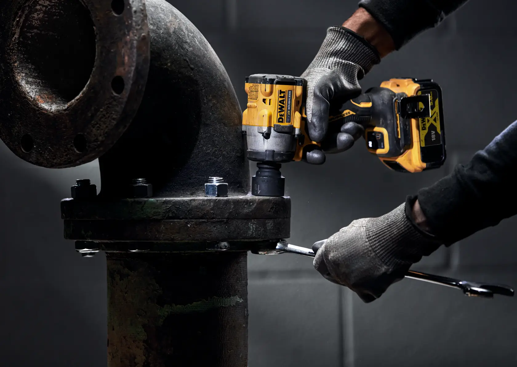 A person wearing gloves uses a DEWALT cordless impact wrench to tighten bolts on a large industrial pipe flange.