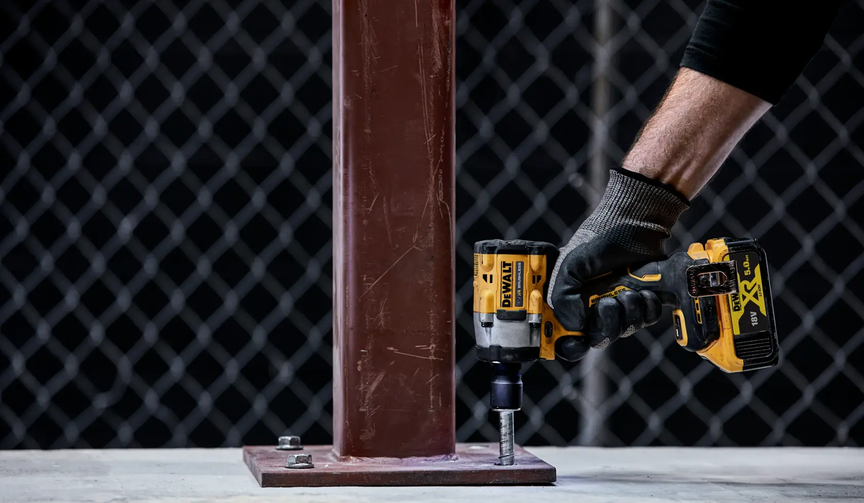 A gloved hand uses a DEWALT cordless impact wrench (model DCF921P2T) to tighten a bolt at the base of a metal column in an industrial setting.