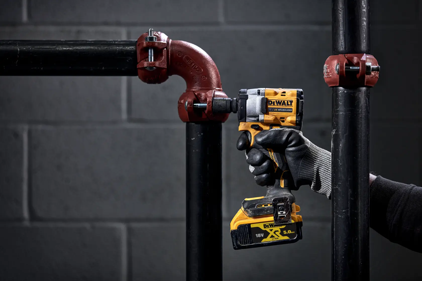 A person wearing a black and grey work glove is using a DEWALT 18V cordless impact wrench on a black metal pipe fitting in an industrial setting. The background is a grey brick wall.