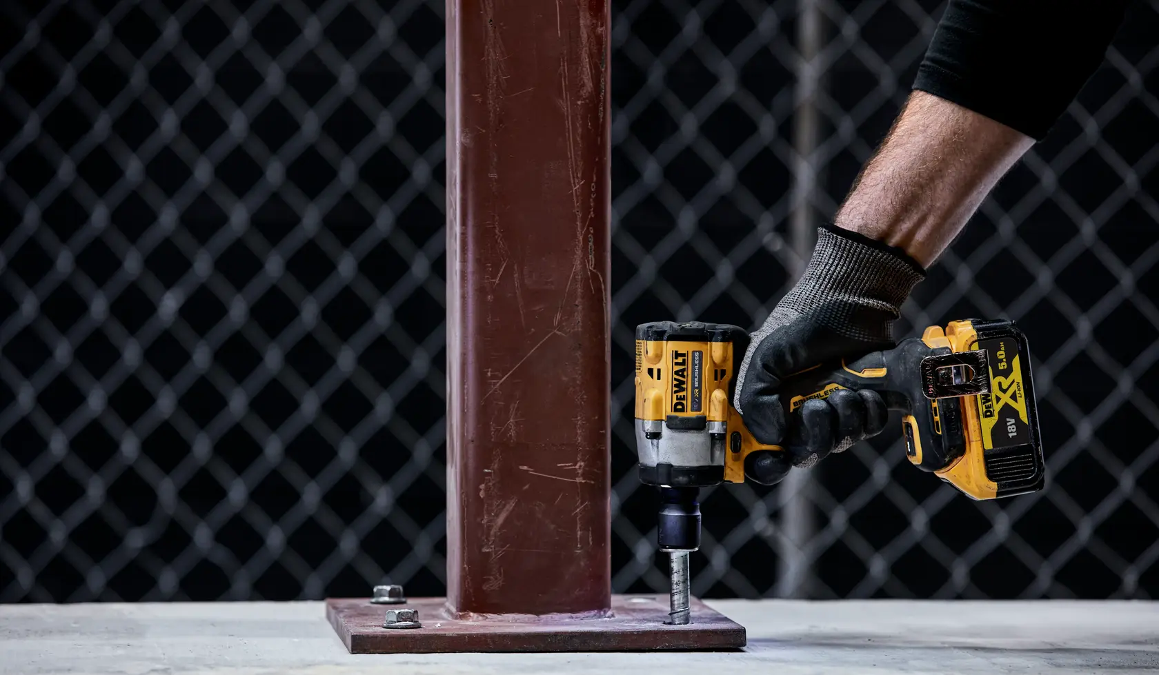 A person wearing a glove is using a DEWALT cordless impact wrench to tighten a bolt on a metal base plate attached to a vertical steel beam.