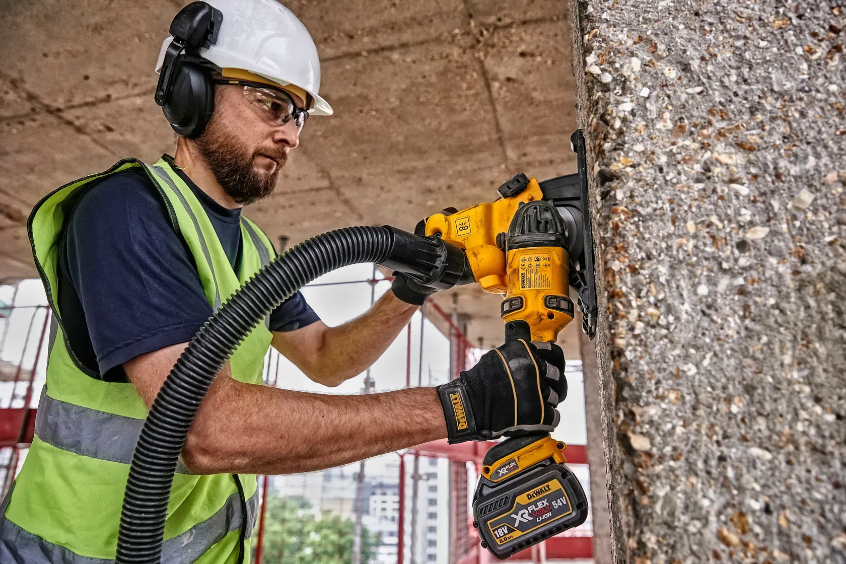A worker wearing a safety helmet, ear protection, and high-visibility vest uses a DEWALT DCG200T2 cordless power tool for grinding concrete on a construction site. The worker's face is blurred for privacy.